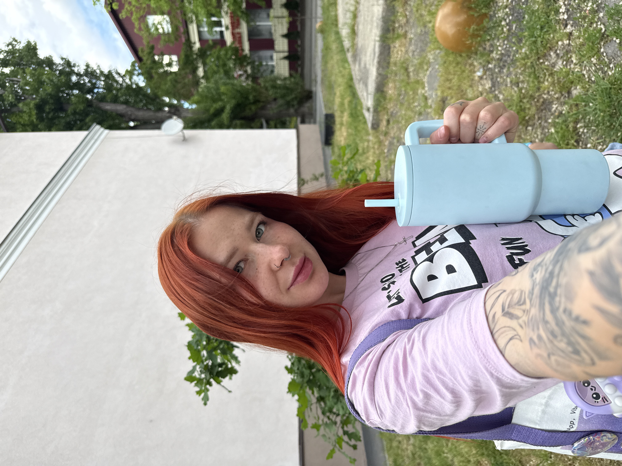 A person with auburn hair and green eyes is holding a light blue cylindrical container with a straw and handle in a selfie taken outdoors. The person is wearing a light purple long-sleeve shirt with bold, graphic black and white lettering. Tattoos are visible on the person's hand and arm. In the background, there's a green lawn, trees, a white building, and a red and white building. An orange round object sits on the lawn near a concrete walkway. The location is possibly in Rostov-on-Don, Russia.