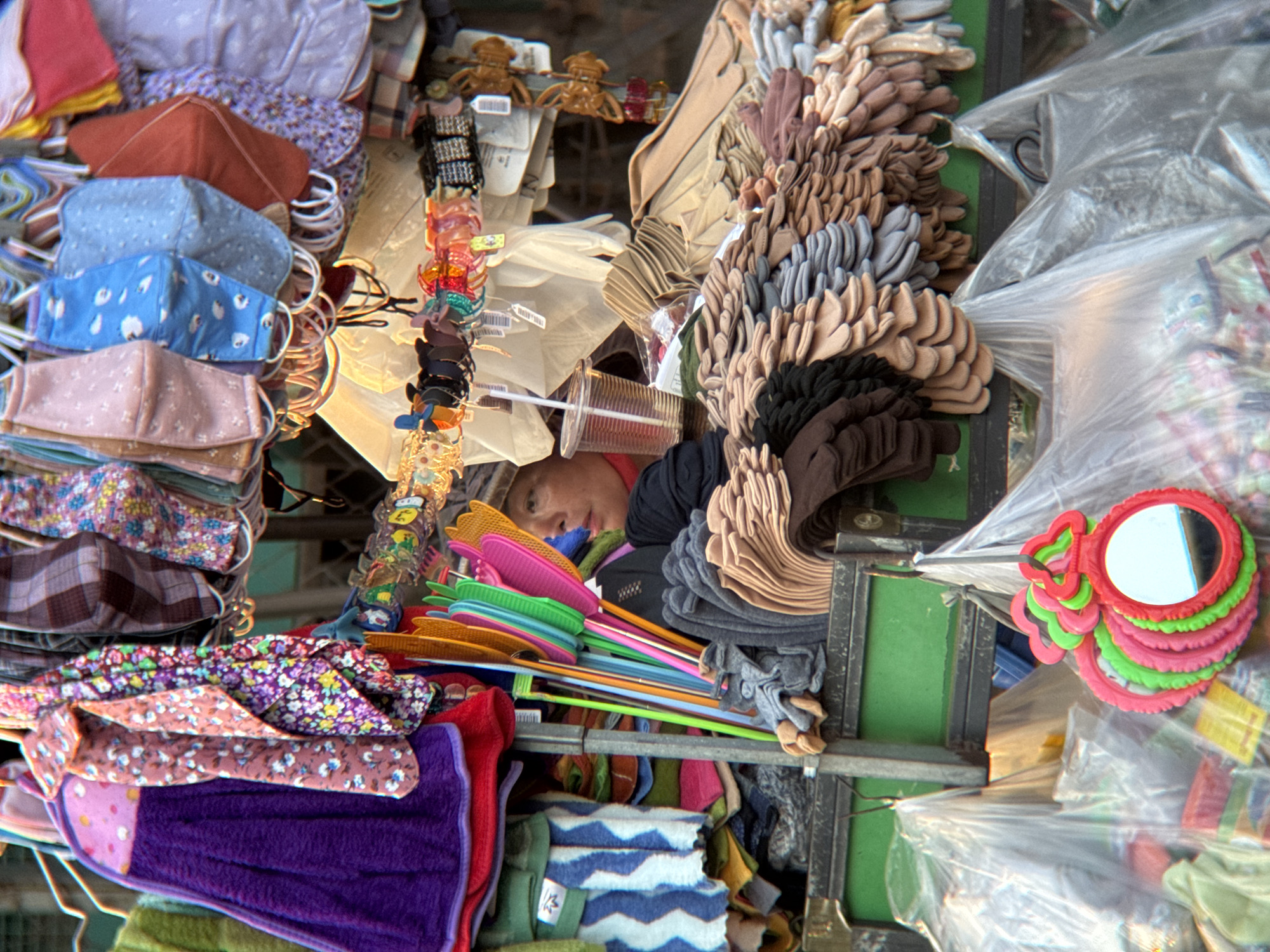This is a street market scene in Nha Trang, Vietnam, likely a roadside stall. A person, appearing to be of Asian descent with dark hair and wearing a dark shirt and a cap, is visible behind a display of goods. They are looking to their right, with their head tilted slightly down and a plastic cup with a straw in front of them.
The stall is packed with various items. In the foreground, there are many pairs of gloves neatly stacked, displayed in rows of different colors, including beige, grey, and brown. Above the gloves, colorful plastic handheld fans are arranged vertically. Behind these, there are racks of fabric face masks, featuring different patterns and colors, including floral, polka dot, and solid blue designs. There are also various other textiles and accessories visible, such as a purple towel or cloth draped over the stall, and a selection of colorful hair clips or barrettes hanging from a rack. Plastic bags filled with unseen items are hanging from hooks below the stall. The overall impression is of a busy, diverse market stall selling a wide array of small consumer goods. The lighting suggests it is daytime, possibly late afternoon, with some shadows present.