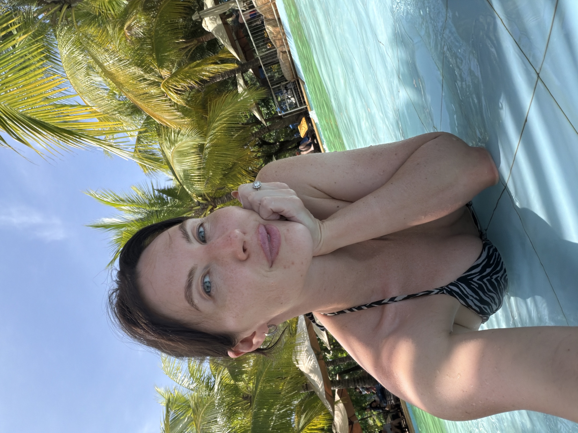 A woman with dark hair and blue eyes is posing for a selfie in a swimming pool. She is wearing a black and white zebra-striped bikini top and a ring on her left hand. Her left arm is resting on her chin, and she is smiling. The pool is filled with clear blue water, and palm trees are visible in the background. The scene appears to be set in a tropical location, likely a resort or hotel. While the specific location in Tay Nha Trang Ward, Vietnam is not definitively identifiable from the image alone, the presence of palm trees and a resort-style pool suggests a vacation destination within that region.