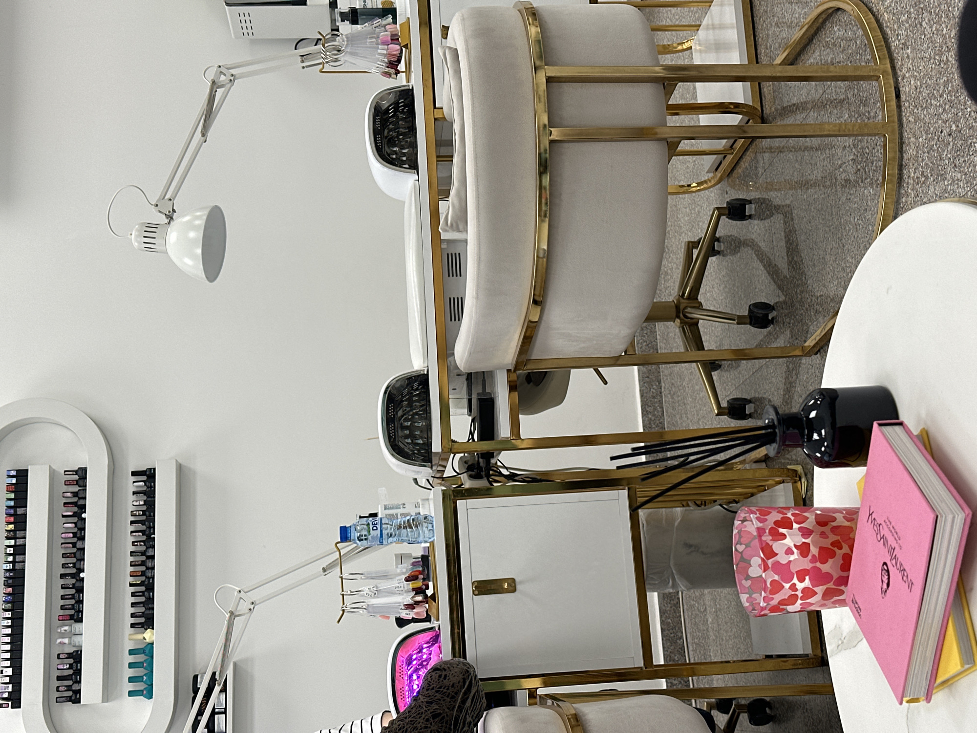 The image depicts a well-lit interior, possibly a beauty salon, in Sofia, Bulgaria. The scene features two manicure stations, each with a white chair, a gold-framed workstation, a white adjustable lamp, and a UV nail lamp. One station is occupied by a person wearing a striped top, with their hand under the UV lamp. The second station is unoccupied. A white shelf displays a variety of nail polish colors. A small white table holds a black diffuser, a candle with red hearts, and a stack of books, one with a pink cover bearing the name "YVES SAINT LAURENT". The background is plain white. The overall impression is a clean and modern aesthetic.