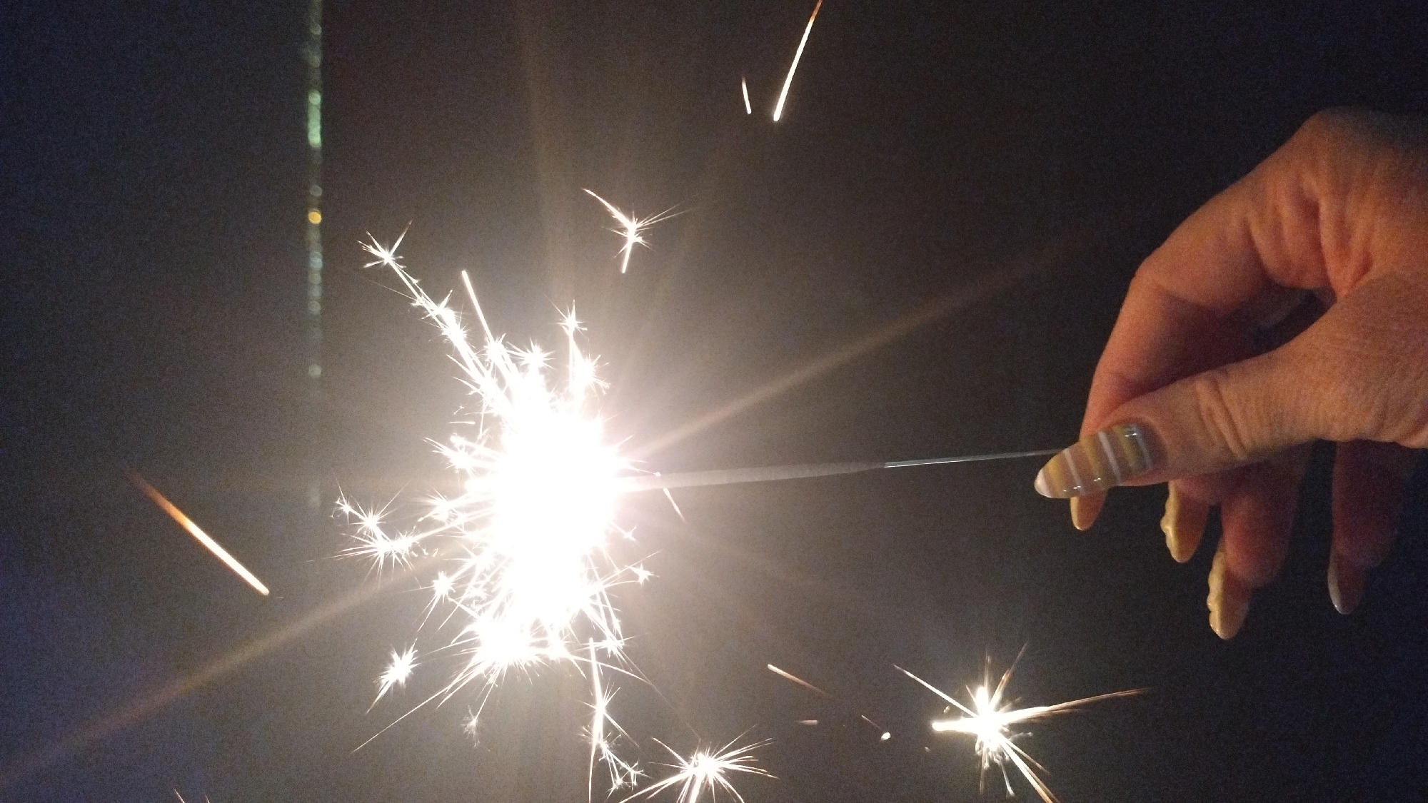 A hand with a striped fingernail design is holding a lit sparkler in front of a dark background. The sparkler is emitting bright white sparks in a starburst pattern. Other sparks are scattered in the background, with some appearing as short, orange lines. In the distance, a blurred line of lights suggests a city skyline. The scene appears to be outdoors at night. There is no specific information in the image to determine its location in Perea, Greece.
