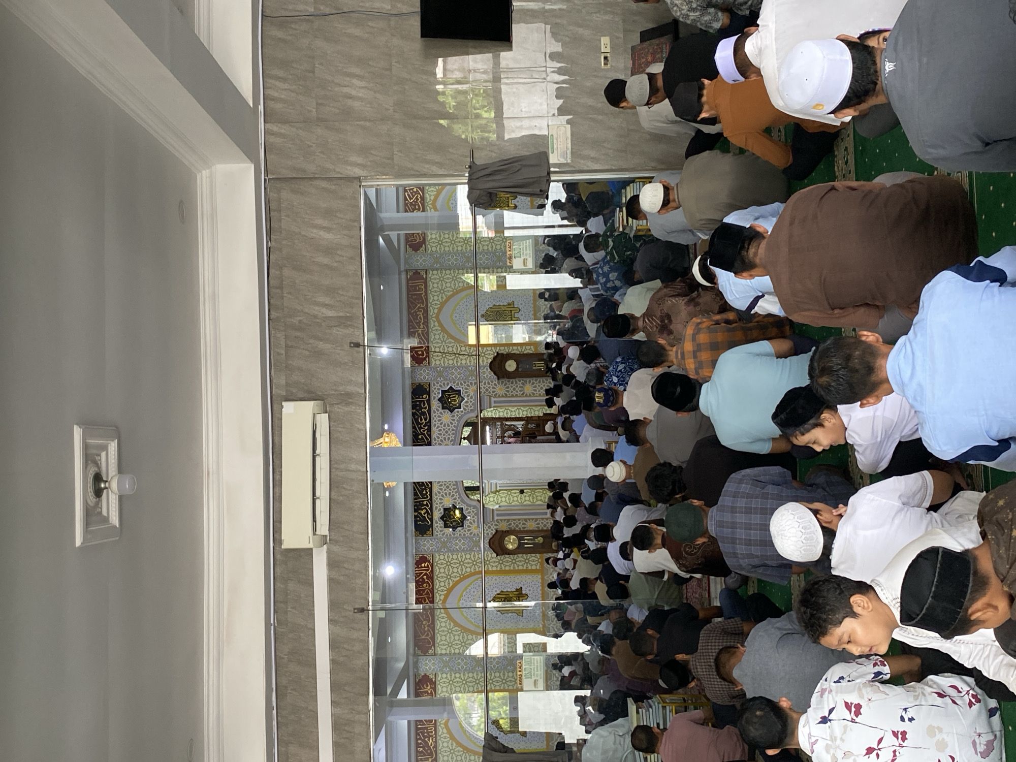 The image depicts a group of Muslim men and boys gathered inside a mosque, likely for prayer, as indicated by their attire and the setting. The mosque appears to be modern, with tiled walls and a reflective glass partition revealing the prayer hall behind it. The prayer hall itself features traditional Islamic architectural elements, including arched doorways and ornate calligraphy on the walls. Several men are seated on a green carpeted floor, facing the direction of prayer. Some are wearing prayer caps, while others have their heads uncovered. The overall atmosphere suggests a communal religious gathering. The location is identified as Lhokseumawe, Indonesia.