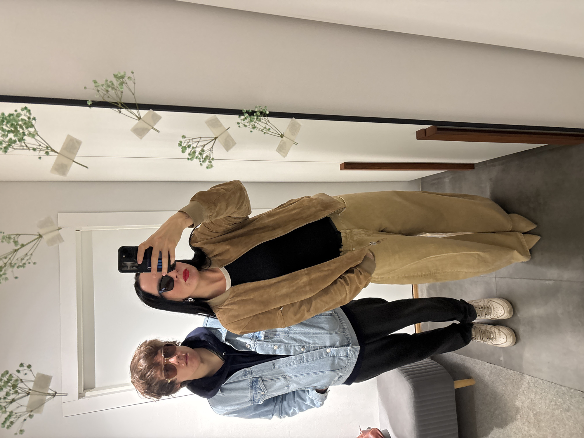 An indoor mirror selfie, taken in Chaikovsky, Russia, features a woman and a man. The woman is in the foreground, holding a black smartphone with a blue-striped case to take the picture. She has dark hair, wears sunglasses, prominent red lipstick, a black top, and a light brown bomber jacket, paired with beige wide-leg pants. The man stands behind her, wearing sunglasses, a dark hoodie under a light blue denim jacket, black pants, and white sneakers.
The setting is a minimalist room, possibly a dressing room or retail space, with a large mirror dominating the scene. Delicate green plant sprigs, resembling baby's breath, are decoratively taped to the mirror at various points. In the upper background, dark wooden beams are visible against white walls. The floor is a neutral light gray, and a gray upholstered bench or seat is partially visible in the bottom right corner. The overall impression is casual and stylish. No discernible text is visible beyond the design on the phone case.