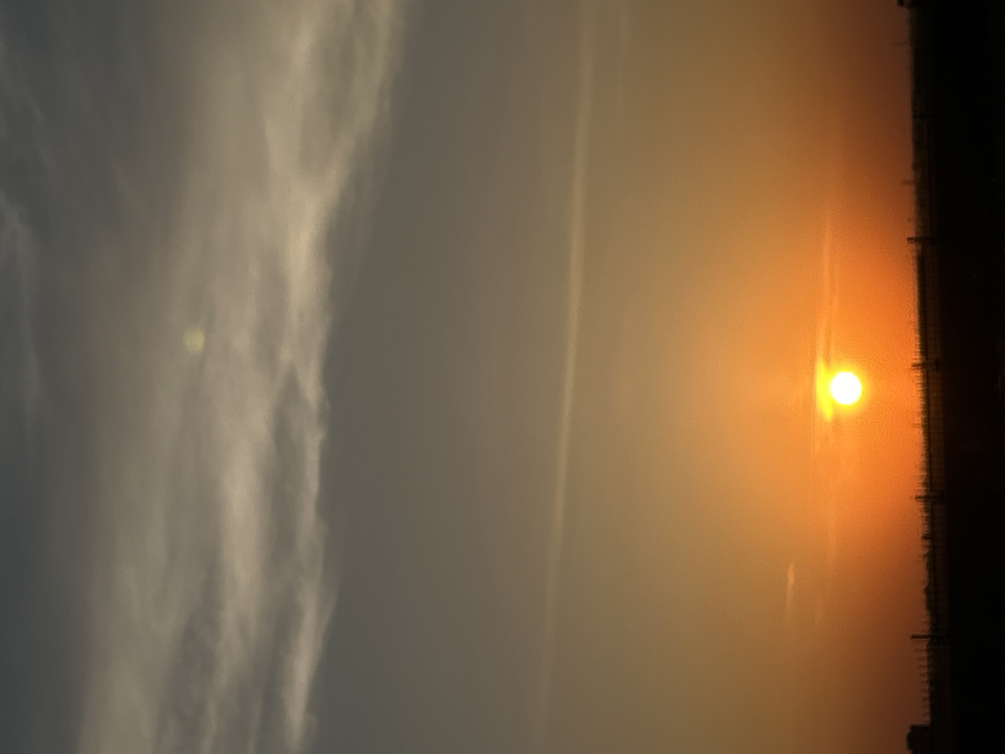 The image shows a sunset over a cityscape. The sun is a bright yellow orb, positioned near the horizon. The sky is filled with orange and yellow hues that transition to grey to the left of the image. The silhouette of buildings is visible along the horizon. It is impossible to tell where in the city of Unknown, Unknown this scene is.
