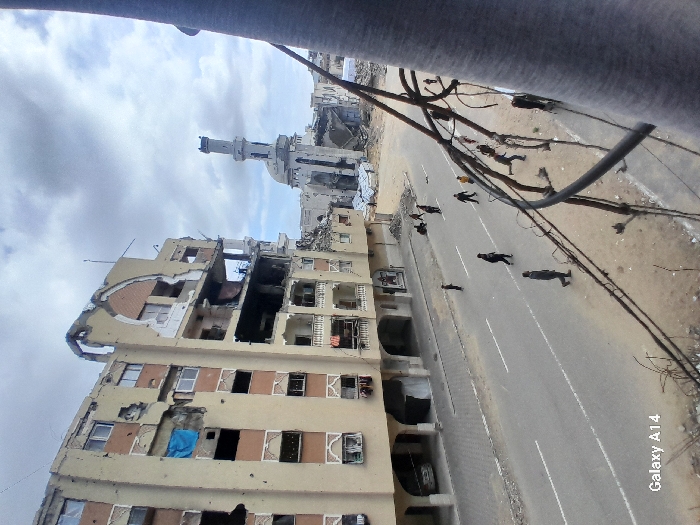 The image depicts a street scene in Khan Yunis, Palestinian Territory, characterized by widespread structural damage under an overcast sky. In the foreground, numerous black electrical wires and a partial view of a grey structure on the right frame the scene. To the left, a multi-story, light-colored residential building shows extensive damage, with multiple floors exposed and walls partially collapsed. A blue tarp is visible in an upper-floor window, and laundry hangs from some balconies. A white vehicle is parked at ground level under an archway.
On the asphalt street below, marked with white lines, approximately 10-15 individuals, appearing to be civilians, walk or stand. Some are on the paved sidewalk adjacent to the damaged building, while others are distributed across the roadway. In the midground and background, further destruction is evident. A prominent mosque with a tall minaret and a dome stands damaged, its dome partially collapsed and walls showing significant impact. Piles of rubble and debris are scattered throughout the background, indicating severe devastation. The overall scene suggests a post-conflict environment.