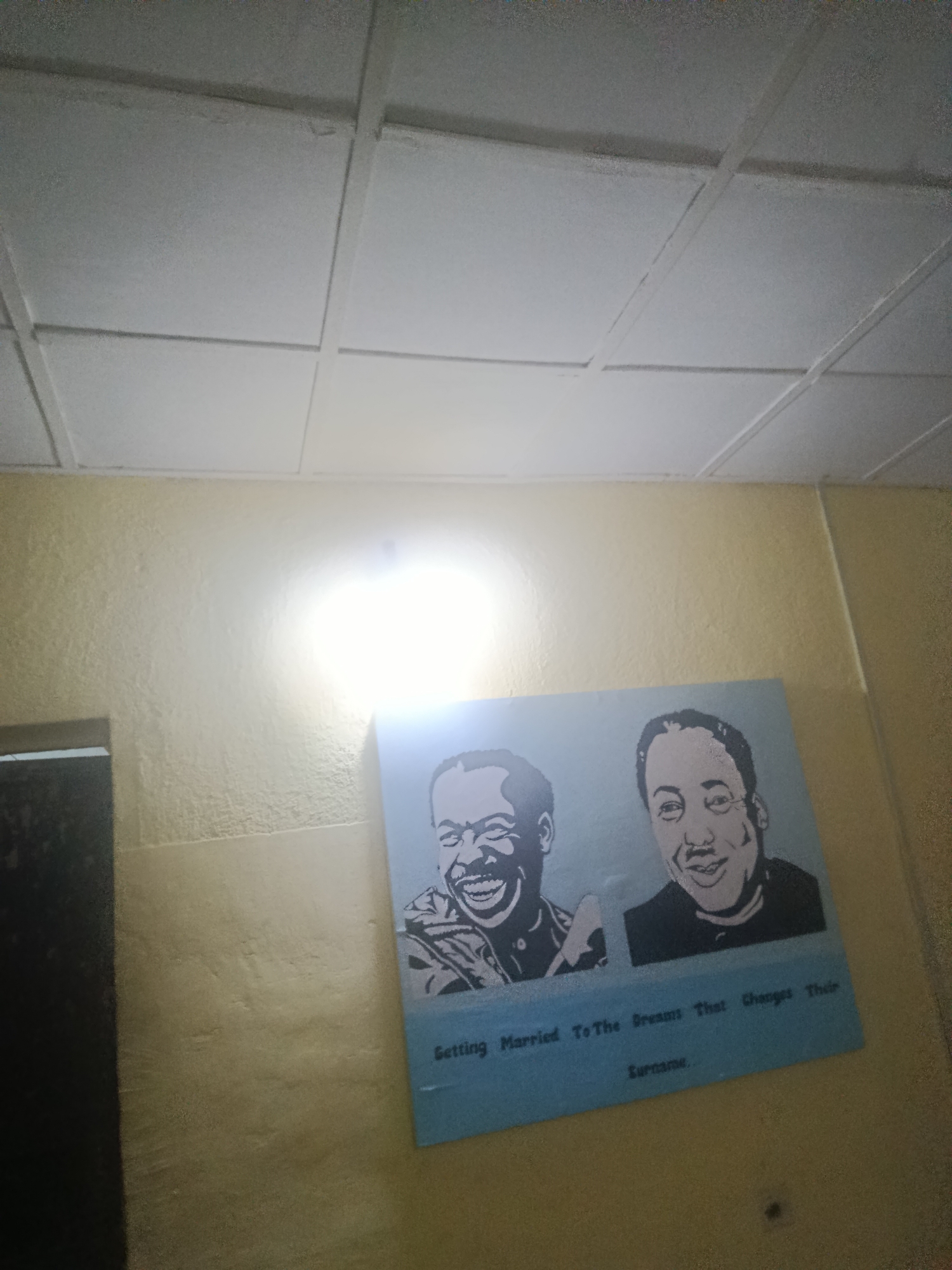 The image shows a low-angle view of an interior wall in Barakin Lerawi, Nigeria. The wall, painted in a light yellow or beige hue, has an uneven texture. A light source casts a bright glare on the wall. To the left, part of a dark, vertical object is visible. On the wall, there's a blue rectangular sign displaying black and white portraits of two men. Beneath the portraits, printed text reads "Getting Married To The Dreams That Changes Their Surname." Above the wall is a white ceiling with a grid pattern.