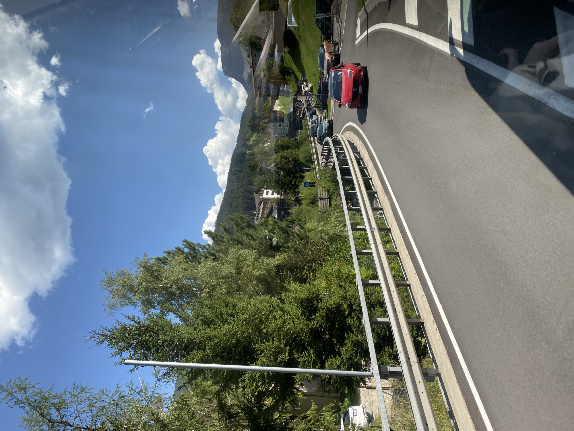 The image depicts a scenic view of a winding road in the mountainous region of Urtijëi - St. Ulrich in Gröden - Ortisei, Italy. The road, with a metal guardrail on the left side, curves to the right, leading towards a cluster of buildings nestled on a hillside. Several cars are visible on the road, including a prominent red car in the foreground and other vehicles further ahead. Dense green trees line the left side of the road, with more foliage and buildings visible on the slopes behind. The sky above is blue with scattered white clouds, and a mountain peak can be seen in the distance. A white vehicle is parked on the left side of the road, beyond the guardrail. The overall scene conveys a sense of travel through a picturesque, possibly resort-like, mountainous environment.