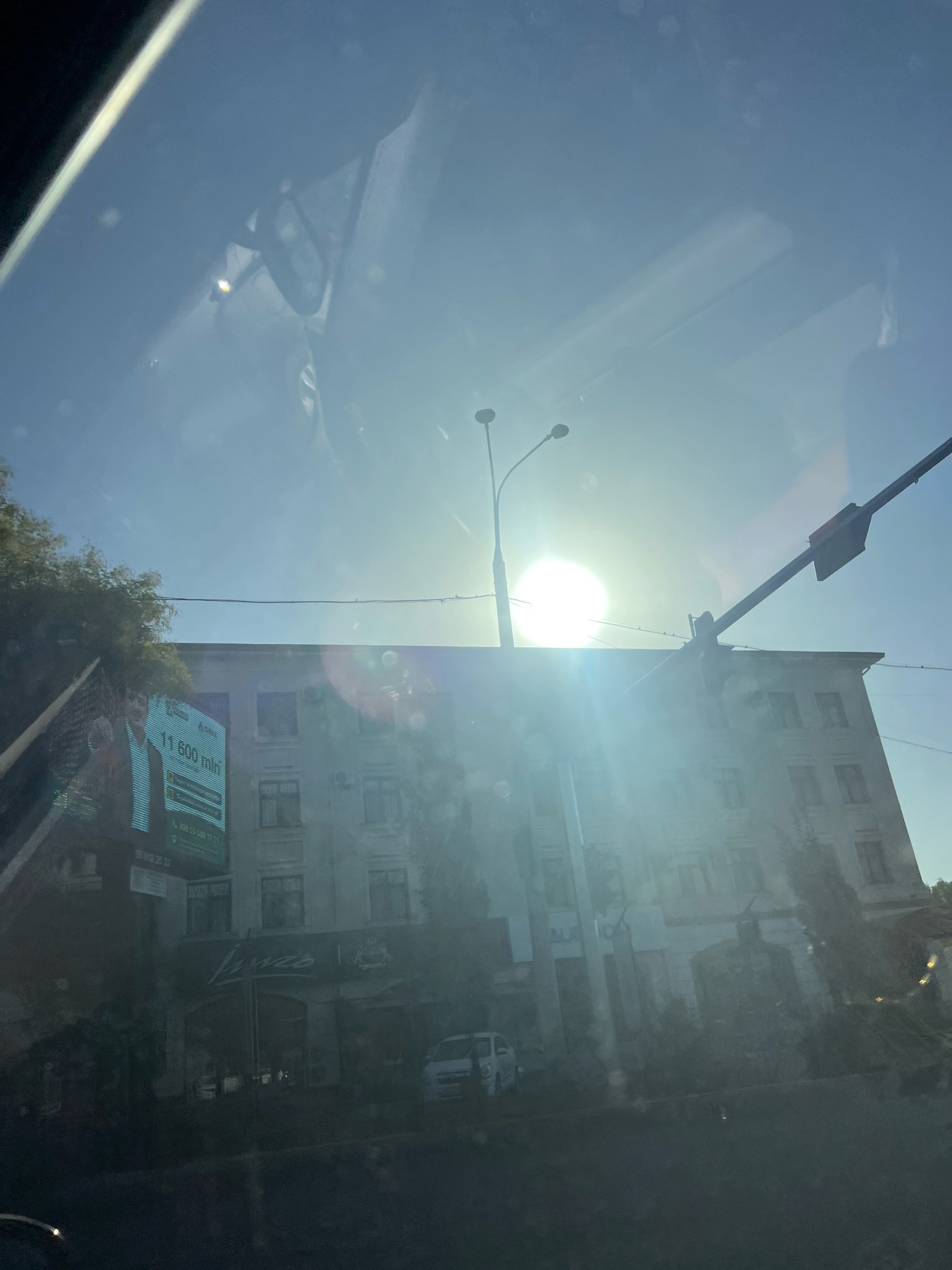 The image shows a sun-drenched street scene with a building in the background. The sun is very bright, creating a strong glare and lens flare that obscures some details. In the foreground, there are reflections on what appears to be a car windshield.
The building in the background is a multi-story structure with windows. There is a large billboard on the left side of the image advertising "11 600 mln". Below the billboard, on the ground floor of the building, is a business with the name "Lusso" visible above an arched entrance. A white van is parked to the right of the "Lusso" establishment.
The environment appears to be urban, with a street and trees visible. A lamppost with two lights is also present. The sky is clear and blue, but the bright sun is the dominant feature. The image is taken from inside a vehicle, as indicated by the reflections and the perspective.