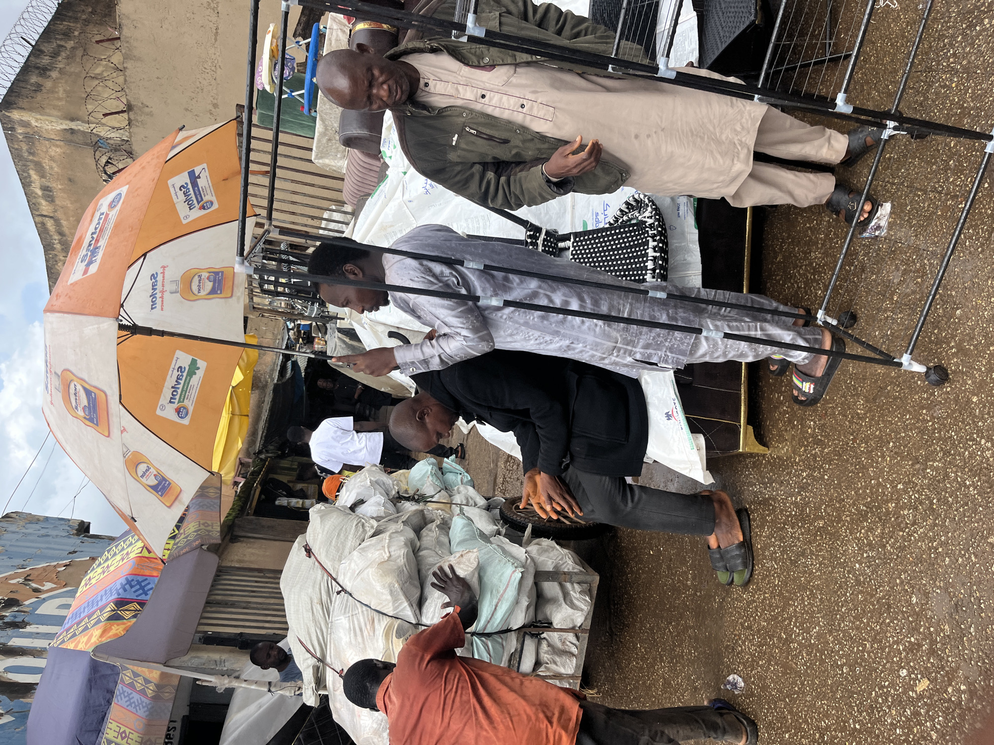 In the city of Jos, Nigeria, a busy outdoor market scene unfolds under a cloudy sky. A large, orange and white promotional umbrella, emblazoned with the "Savlon" brand, provides shade. Several individuals are present. In the foreground, a man in a green jacket and tan traditional attire stands near a metal rack. To his left, another man in a silver tunic is looking at his phone. Behind them, a cart overflowing with large, white sacks is being pushed by a man in an orange shirt. Another man, dressed in a black coat and dark pants, sits on a low structure, his hands clasped. The ground is wet and reflective, suggesting recent rain. The market appears to be a place of commerce, with goods piled high and people engaged in various activities.