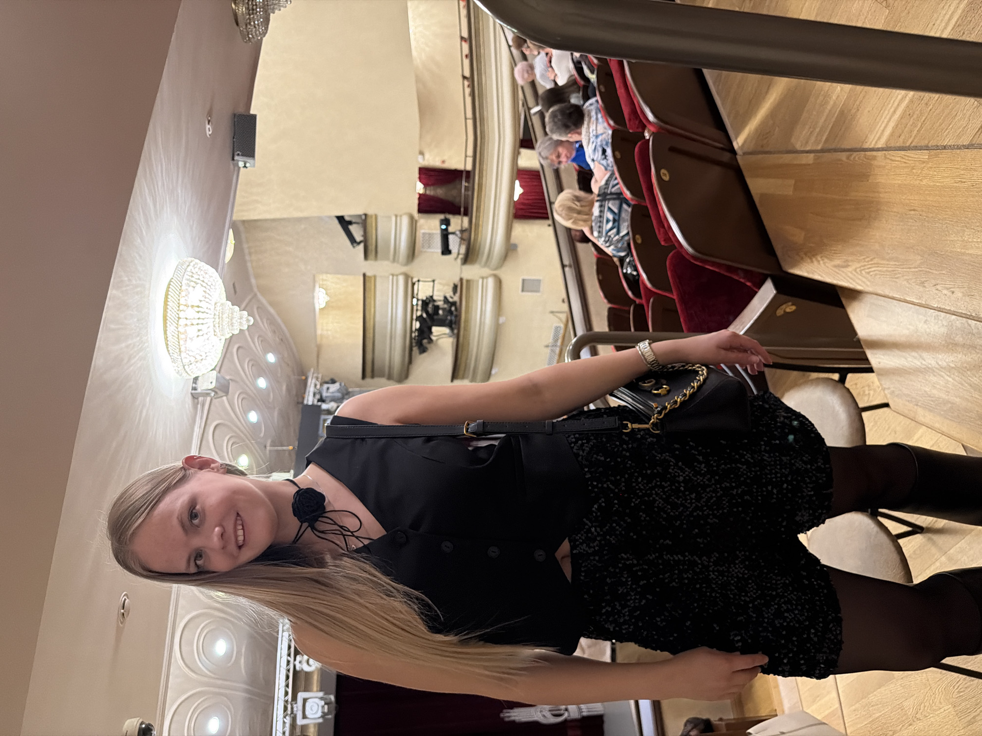 The image shows a woman posing inside what appears to be a theater. The architectural design and ornate detailing visible in the background, particularly the elaborate chandeliers and patterned walls, suggest a grand, possibly historic interior. Rows of seated theatergoers can be seen in the background, indicating that the venue may be in use or prepared for an event. The woman is wearing a black outfit with boots. The presence of theatergoers, the architectural grandeur, and the woman's attire suggest a social or entertainment setting. It is unknown if the image was taken in Izhevsk, Russia.
