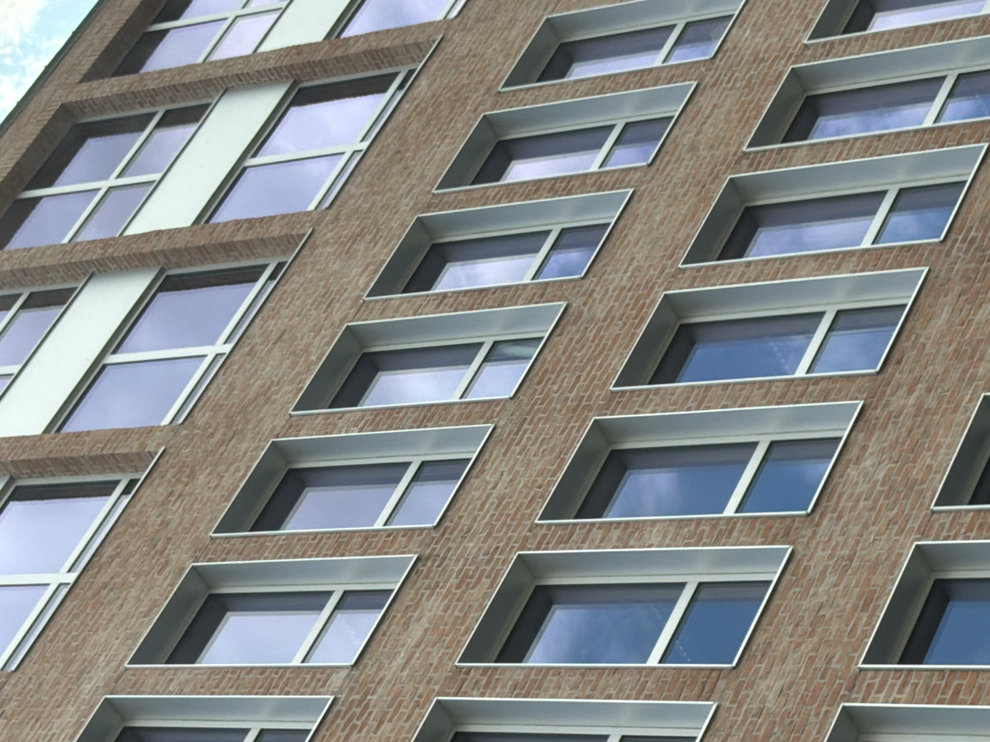 This media file showcases the facade of a modern building, characterized by its repetitive arrangement of windows set into a brick exterior. The windows are rectangular and appear to be made of glass with white frames. They are set at various angles and depths, creating a visually dynamic pattern across the building's surface. Some windows are flush with the brickwork, while others are recessed, forming small, angled ledges. The building's facade is constructed from reddish-brown bricks, laid in a regular pattern. Above the windows, there are horizontal bands of a lighter material, possibly metal or concrete, that provide a visual separation between rows of windows. The sky is visible in the reflections of the glass, suggesting it is daytime. The lighting indicates a bright but not harsh sunlight, and the clear sky in the reflections suggests fair weather. The overall impression is one of contemporary architecture, with a focus on geometric design and the interplay of light and shadow on the building's surface. The scene is an exterior architectural view, likely in an urban setting given the building's design. There is no visible text in the image.