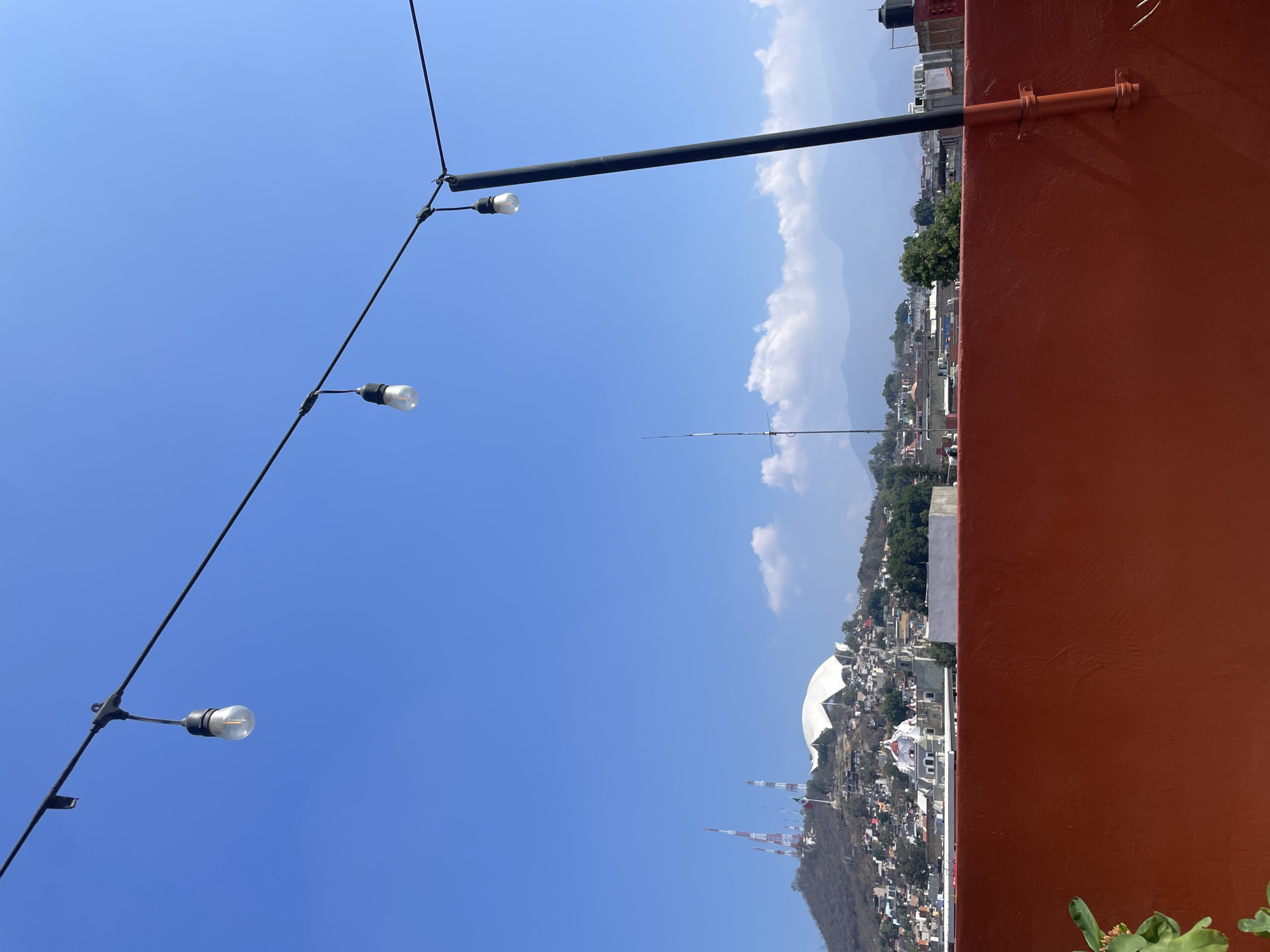 The image depicts a rooftop view in Oaxaca City, Mexico, on a clear, sunny day. The sky is predominantly blue with some wispy white clouds in the distance, near the mountain range.
In the foreground, there's a dark brown-red wall occupying the right side of the frame. A black metal pole with string lights is visible, extending from the left side of the frame and diagonally across the scene. The lights appear to be clear glass bulbs.
In the middle ground, the city sprawls with densely packed buildings, including the dome-shaped Auditorio Guelaguetza. There are radio or cell towers, and a sharp, thin spire rises from the landscape. The environment includes a mix of residential and commercial structures.