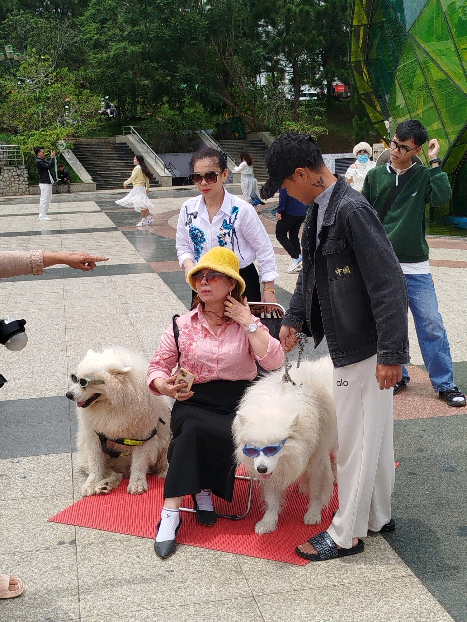 In a public outdoor plaza in Da Lat, Vietnam, likely near a tourist attraction given the modern architectural structure with green, faceted glass visible in the background, a group of people and two Samoyed dogs are gathered. In the foreground, a woman wearing a pink collared shirt, a black skirt, a yellow bucket hat, and sunglasses is seated on a red mat, holding a smartphone and touching her face. Next to her sits a Samoyed dog wearing sunglasses and a harness. Another Samoyed dog, also wearing sunglasses, stands beside her. To the right, a man in a black denim jacket, white pants with "alo" written on the leg, and a tattoo on his neck, holds the leash of the standing dog. He is looking towards the right of the frame. Behind them, other individuals are visible, including a woman in a white blouse with blue embroidery and sunglasses, and a man in a green hoodie and glasses. In the middle ground, more people are seen walking, including a woman in a white dress twirling. The ground is paved with checkered tiles. Steps and greenery are visible in the background. The overall scene suggests a casual outing or a photo opportunity with the dogs.