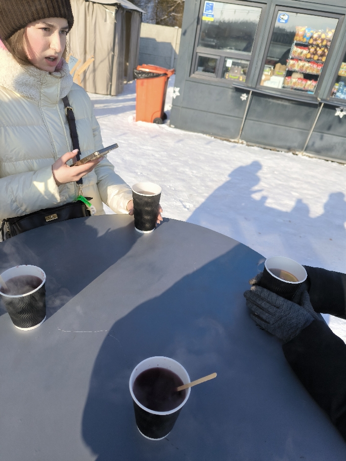 The scene depicts an outdoor gathering during daylight in a snowy environment in Prybuzke, Ukraine. A round, dark grey table occupies the foreground and midground, covered in sunlight and shadows. Four black, corrugated paper cups containing hot beverages are placed on the table; three hold a dark liquid, emitting steam, and one contains a lighter liquid with a lemon slice. A wooden stir stick is present in one of the dark liquid cups.
On the left side of the table, a young female, wearing a brown knitted beanie and a cream-colored puffer jacket with a fur-lined collar, holds a smartphone in her left hand and one of the black paper cups in her right. A black shoulder bag with a gold clasp hangs from her left shoulder. Her mouth is slightly ajar.
On the right side of the table, a second individual's gloved hands, clad in dark grey, are seen holding another black paper cup. The hands are positioned as if warming them on the cup.
The ground surrounding the table is covered in an unbroken layer of white snow, with multiple elongated shadows of people cast upon it, suggesting the presence of at least two individuals, possibly more. In the background, an orange wheeled garbage bin with a black liner stands on the snow, adjacent to a dark grey commercial kiosk or stand with windows displaying packaged food items and drinks. Behind the bin, a light-colored, tent-like structure or shed is visible, with bare trees and a fence beyond it. The sky is clear, indicating bright sun conditions.