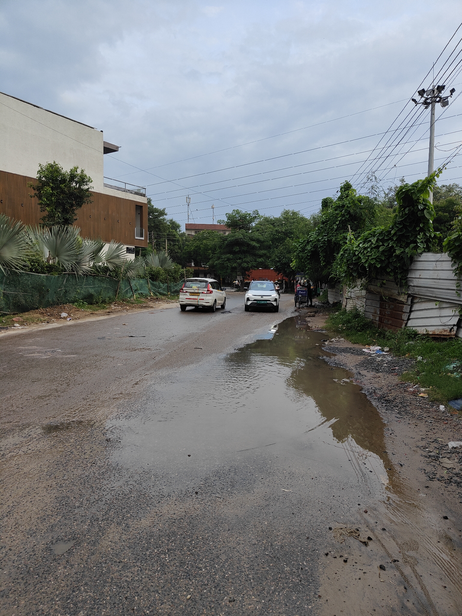 A wide, overhead shot shows a street in Gurgaon, India, with a large puddle reflecting the sky, power lines, and trees. The street is wet and appears to be in poor condition, with visible potholes. On the left side of the road, a white car with an Indian license plate is driving away from the camera. Further down the road, another white car is approaching. On the right side, a man is pushing a cart with the "Amul" logo on it. Buildings and vegetation are visible on both sides of the street, with a modern building with wooden paneling on the left. The sky is overcast with gray clouds.