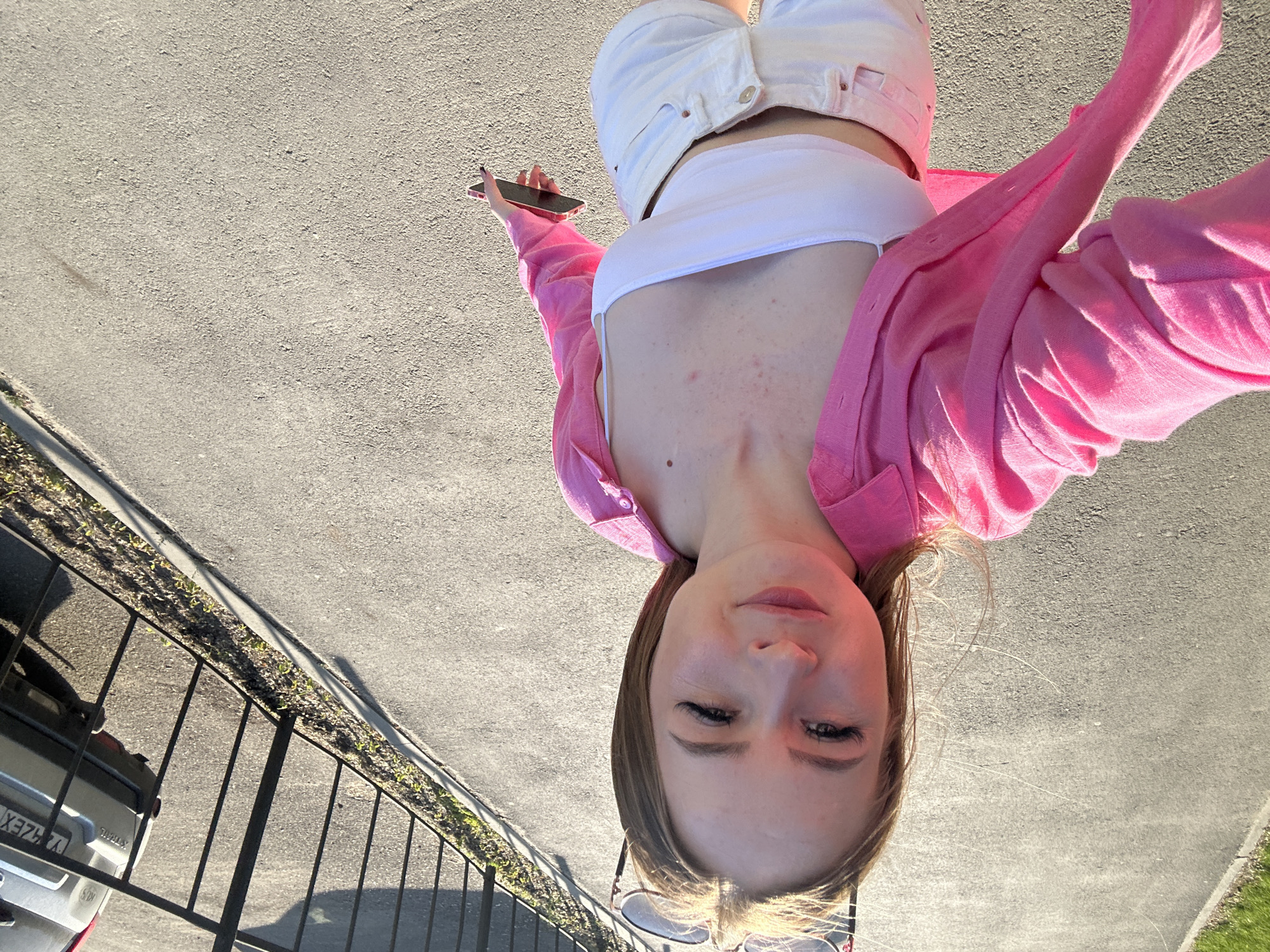 The image, originally oriented vertically with the top of the subject's head pointing right, depicts a young woman taking a selfie outdoors under bright daylight.
The woman is shown from a high-angle perspective, looking down towards the camera she is holding. She has light brown hair and a relaxed expression with a subtle smile. She is wearing a white strapless or bandeau crop top, white high-waisted shorts or trousers, and a bright pink long-sleeved shirt, possibly a cardigan, worn unbuttoned over the crop top. A pair of sunglasses hangs from the neckline of her top. Her right hand, adorned with dark nail polish, holds a smartphone with a red or pink case, positioned to capture the selfie. A small mole and some reddish blemishes are visible on her upper chest.
The setting is an outdoor paved area, likely an asphalt street or parking lot, with strong sunlight casting distinct shadows. To her left, a black metal fence runs alongside a curb, beyond which is a strip of dry grass. Partially visible behind the fence is the rear of a silver car. Text on the car's license plate includes "Y3 4ZEX", and the car model "X-TRAIL" is also identifiable. The overall atmosphere suggests a sunny day.