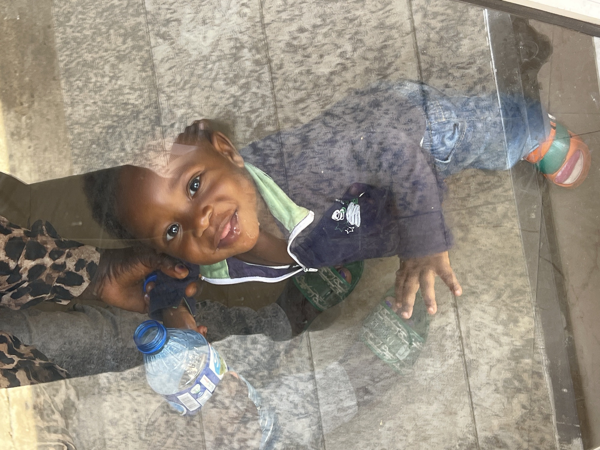A cheerful young child is captured looking up and smiling directly at the viewer through a transparent surface, likely glass or clear plastic. The child, with dark skin and hair, is wearing a navy blue jacket with a light green collar and white zipper, over blue jeans. A bright smile reveals teeth, with a hint of moisture around the mouth, conveying a joyful and engaging emotion. Green sandals are visible on the child's feet, and what appears to be an orange and green shoe is on the child's right foot.
The child is lying down on a light-colored, textured floor, which could be concrete or tile. The transparent surface creates reflections that show parts of an adult, including an arm with a leopard print sleeve and a hand holding a plastic water bottle with a blue cap. The bottle is partly filled with water, and its label has some indistinct text. Another adult hand is also visible near the child's left hand, suggesting interaction. The scene appears to be an indoor or covered outdoor area during daylight hours, consistent with general settings in Gura Top, Nigeria. No specific location cues beyond the floor texture are discernible, nor is the weather visible.