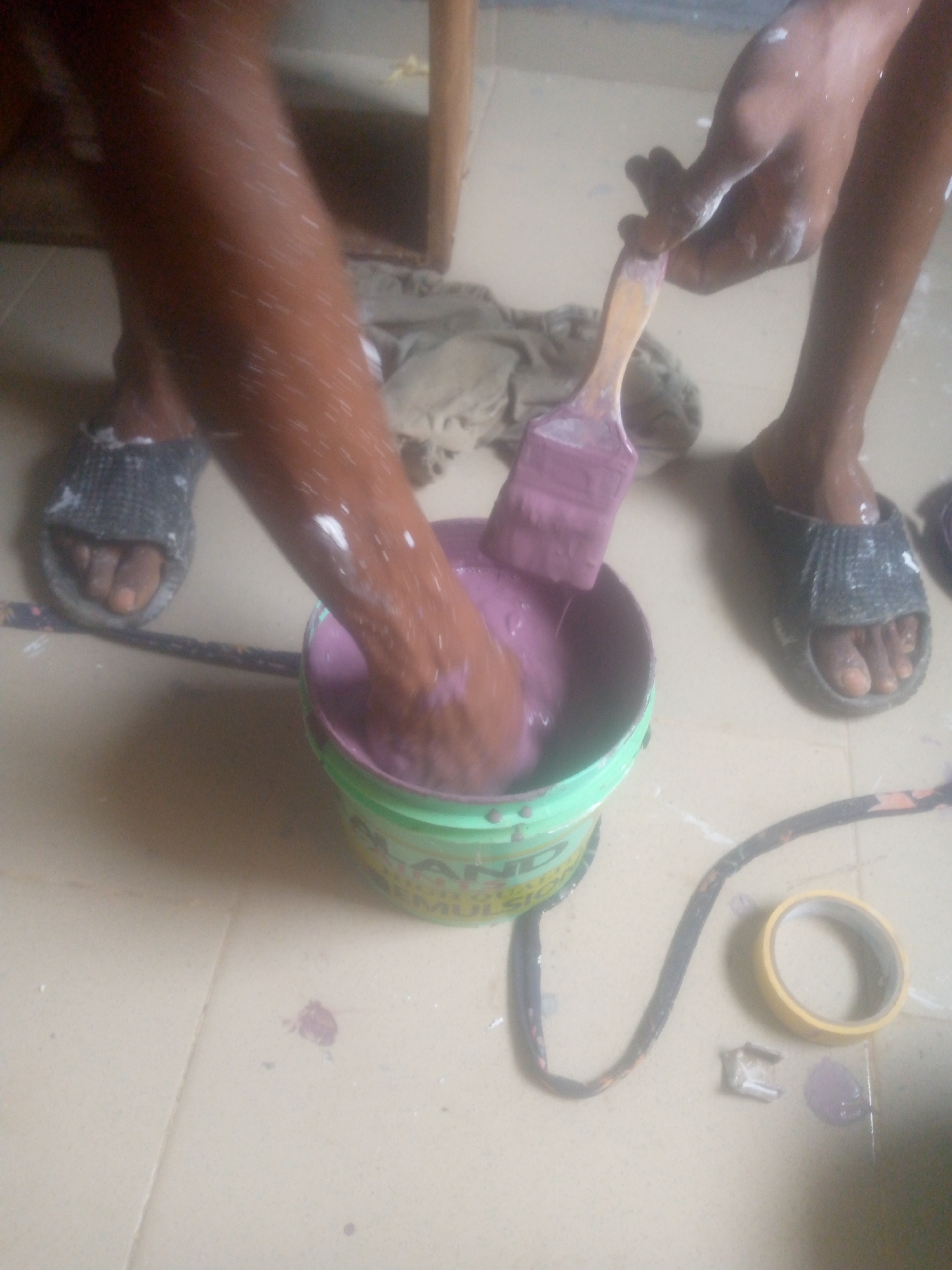 An indoor scene depicts two individuals engaged in a painting activity on a light-colored tiled or concrete floor. At the center is a light green bucket filled with purple paint. One person's right arm, heavily splattered with white and purple paint, is submerged into the paint within the bucket. Their left foot, also bearing paint marks and clad in a dark-colored sandal, is visible to the left of the bucket.
The second person's right hand, also splattered with white paint, holds a wooden-handled paintbrush, with the brush head dipped into the purple paint. This person's right foot, similarly splattered with paint and wearing a dark-colored sandal, is visible to the right. Both individuals are actively involved with the paint, suggesting mixing or preparation.
The floor around the bucket is marked with numerous purple paint splatters. Other objects on the floor include a crumpled piece of light-colored cloth or rag behind the bucket, a black cable or cord (possibly with orange markings) snaking across the floor, a roll of yellow masking tape, and a small, silvery object near the tape. Partial text is visible on the green bucket, including "LAND" and "EMULSI". The setting is a casual indoor workspace, with ambient lighting suggesting daytime. No specific emotions are discernible.