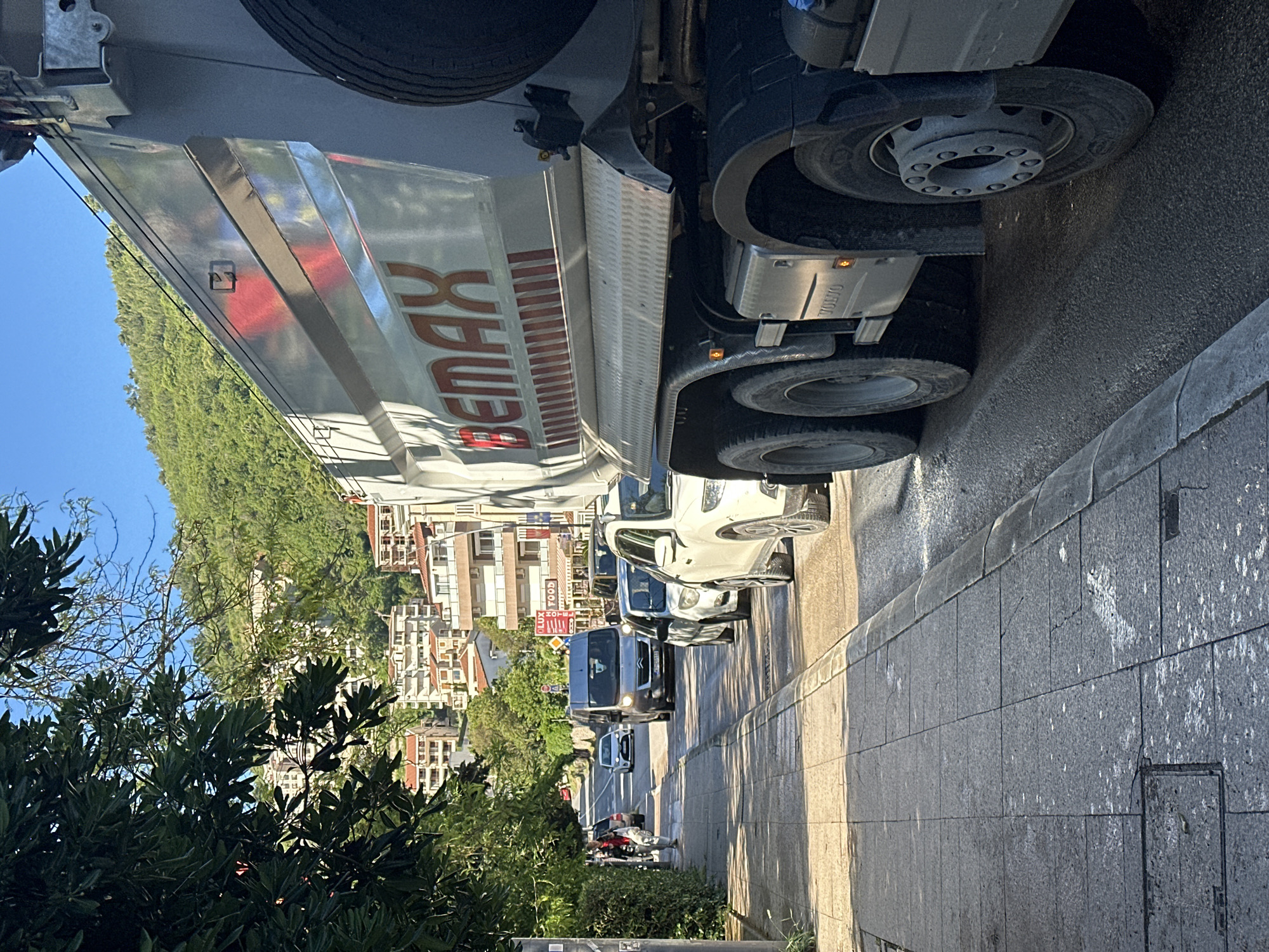 The image depicts a street scene in Budva, Montenegro. A large "BEMAX" branded truck is parked on the right side of the road. Several cars are parked in front of the truck. Pedestrians are on the sidewalk. Buildings and trees are in the background. The sky is blue.