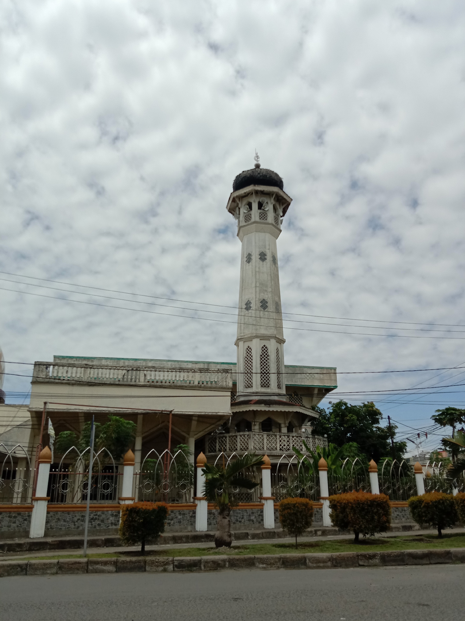 The image shows a mosque in Lhokseumawe, Indonesia. The mosque has a tall, white minaret with a dark, dome-shaped top. The minaret is adorned with geometric patterns. The mosque building is beige and has a flat roof with a balustrade. In front of the mosque, there is a fence with white pillars and curved metal arches, and small, round bushes are planted along the sidewalk. A street is visible in the foreground. Overhead, there are numerous power lines. The sky is overcast with white, puffy clouds.