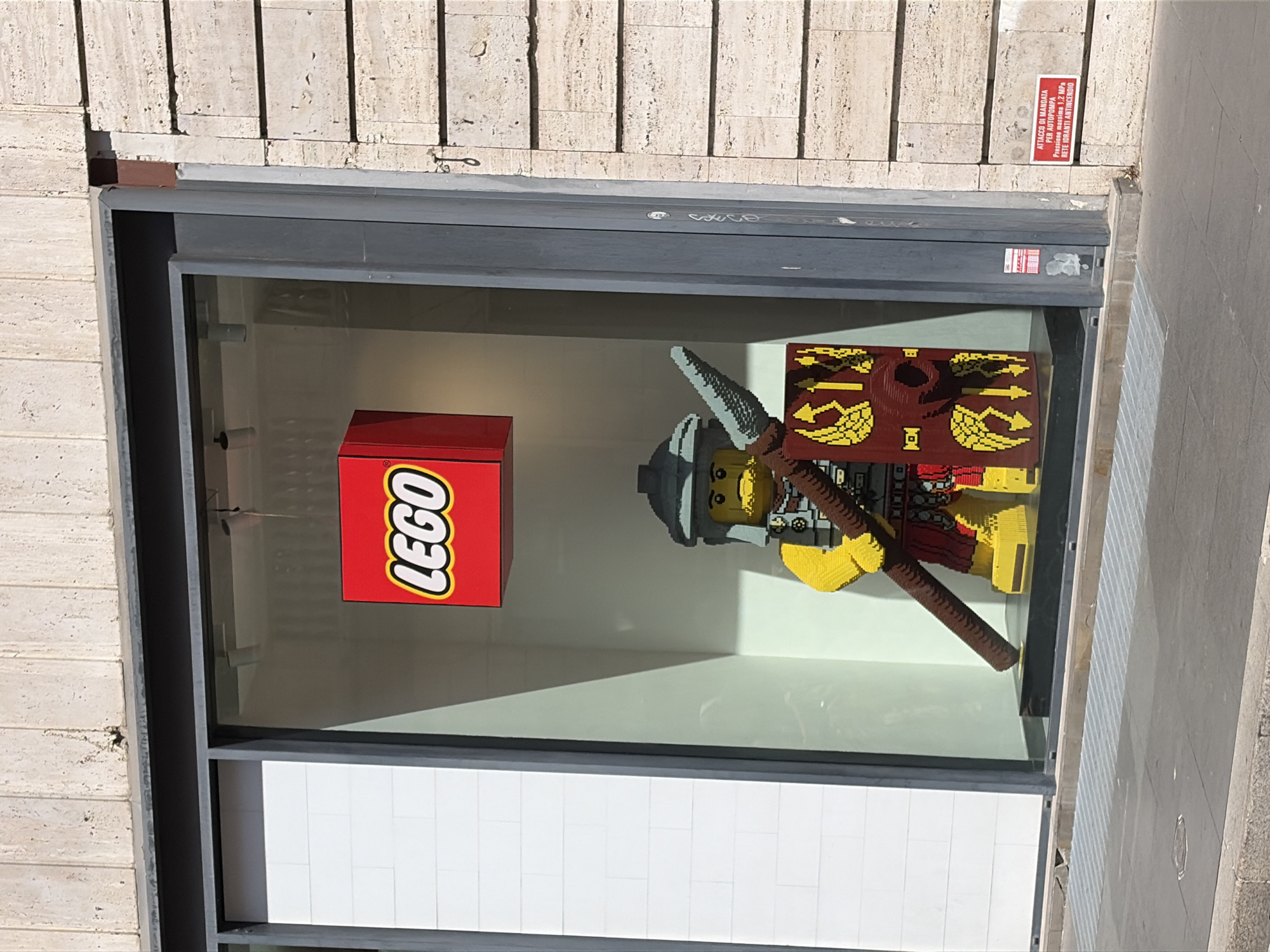 The image shows a Lego store window display featuring a large Lego sculpture of a Roman legionary. The sculpture is life-sized and depicts the figure from the waist up, holding a spear and a shield. The shield is red with yellow wing designs and what appears to be a Roman eagle or wolf symbol. The legionary wears a helmet, armor, and yellow tunic. Above the sculpture, a red Lego logo is prominently displayed. The window itself is large and rectangular, set into a building facade made of light-colored stone. To the left of the display is a white tiled wall. A red sign with Italian text is affixed to the building to the right of the window, which reads "ATTACCO DI MANDATA PER AUTOVìPOMPA Pressione massima 1.2 MPa RETE DURANTE ANTINCENDIO". This indicates a fire hydrant connection. The scene is captured from a street-level perspective, with a sidewalk and a metal grate visible in the foreground. The overall environment suggests a commercial area in Rome, Italy, likely a pedestrian street given the visibility of the sidewalk and grate.