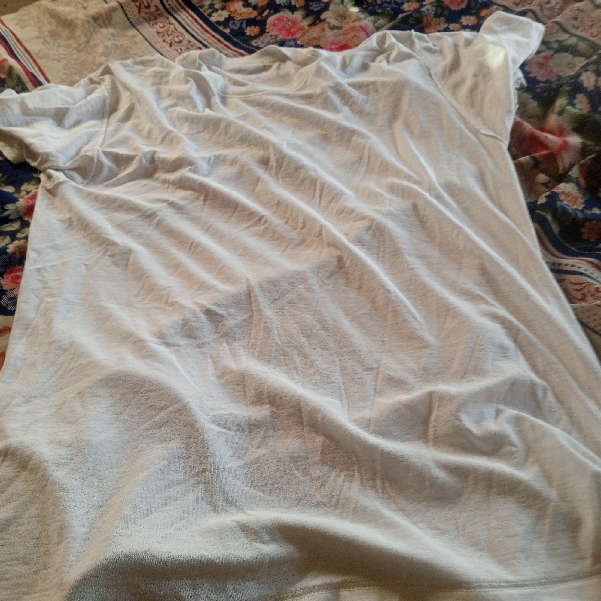 The image displays a crumpled white t-shirt laid out on top of a floral-patterned fabric. The t-shirt is not in its entirety visible, with the top left corner obscured by the floral fabric. The floral fabric itself exhibits a repeating pattern of various flowers, predominantly in shades of pink and blue, against a dark background. The lighting suggests an indoor environment, casting subtle shadows that highlight the folds and texture of the t-shirt. There is no indication of any specific location in Zinna, Nigeria, or any discernible human presence within the frame.