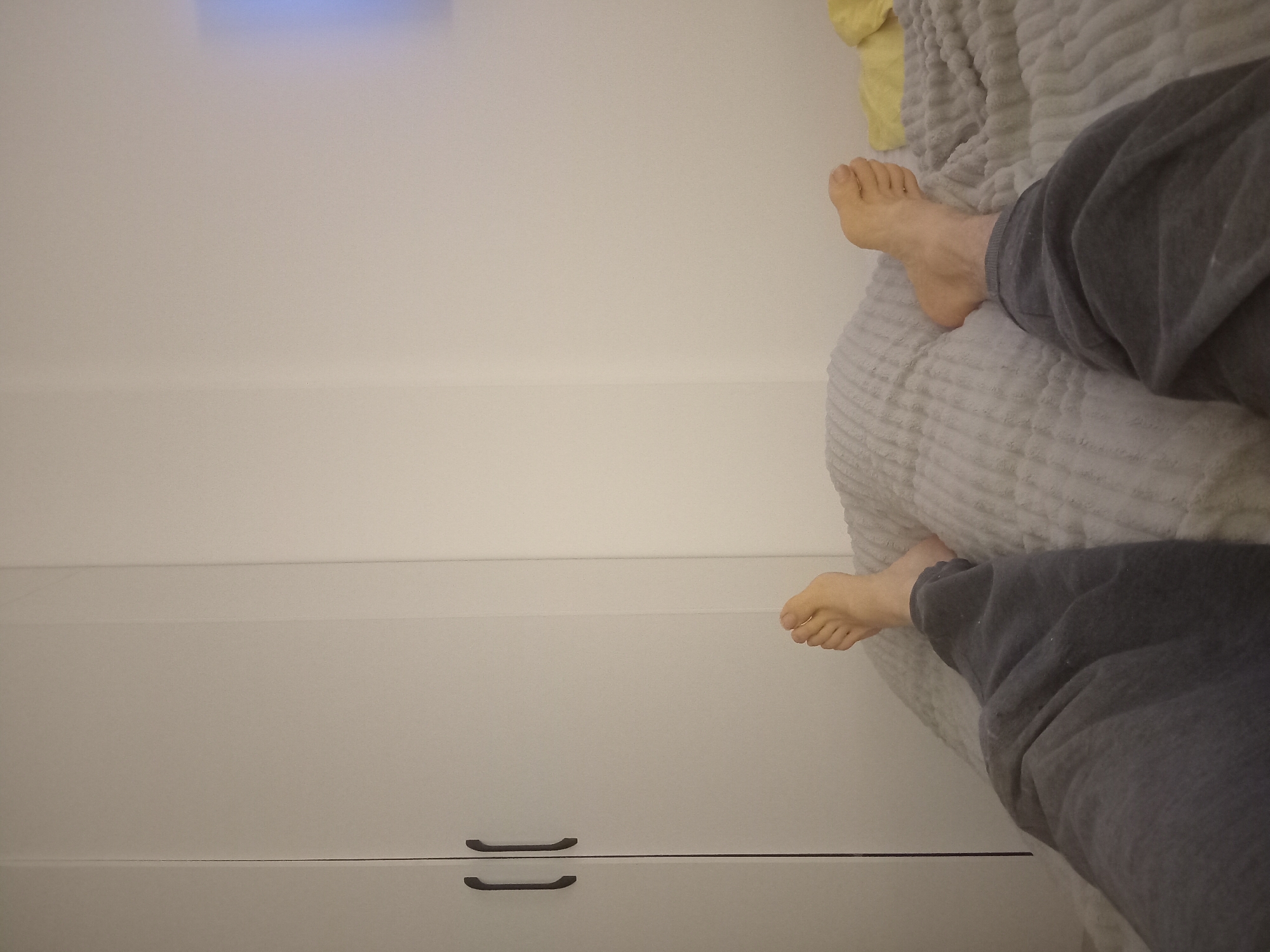 A close-up shot shows a person lying on a bed, with their bare feet extending off the edge of the bed. The person is wearing grey sweatpants. The bed is covered with a textured, light grey blanket. To the left is a white wall, and a set of white drawers with black handles. The location is unknown and unrelated to Batumi, Georgia.