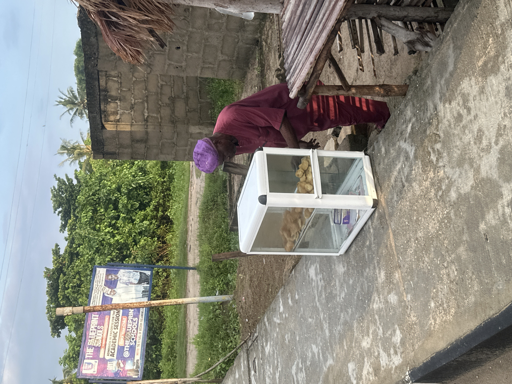 The image shows a man in a maroon traditional outfit and a purple cap standing next to a glass display case filled with baked goods. The display case is white and has two shelves. The man appears to be attending to the goods in the case. In the background, there is a brick building, lush green vegetation, and palm trees. A sign for "THE BLUEPRINT SCHOOLS" is visible on the left side of the image. The scene is outdoors, with a concrete path in the foreground. The location is identified as Badagry, Nigeria.