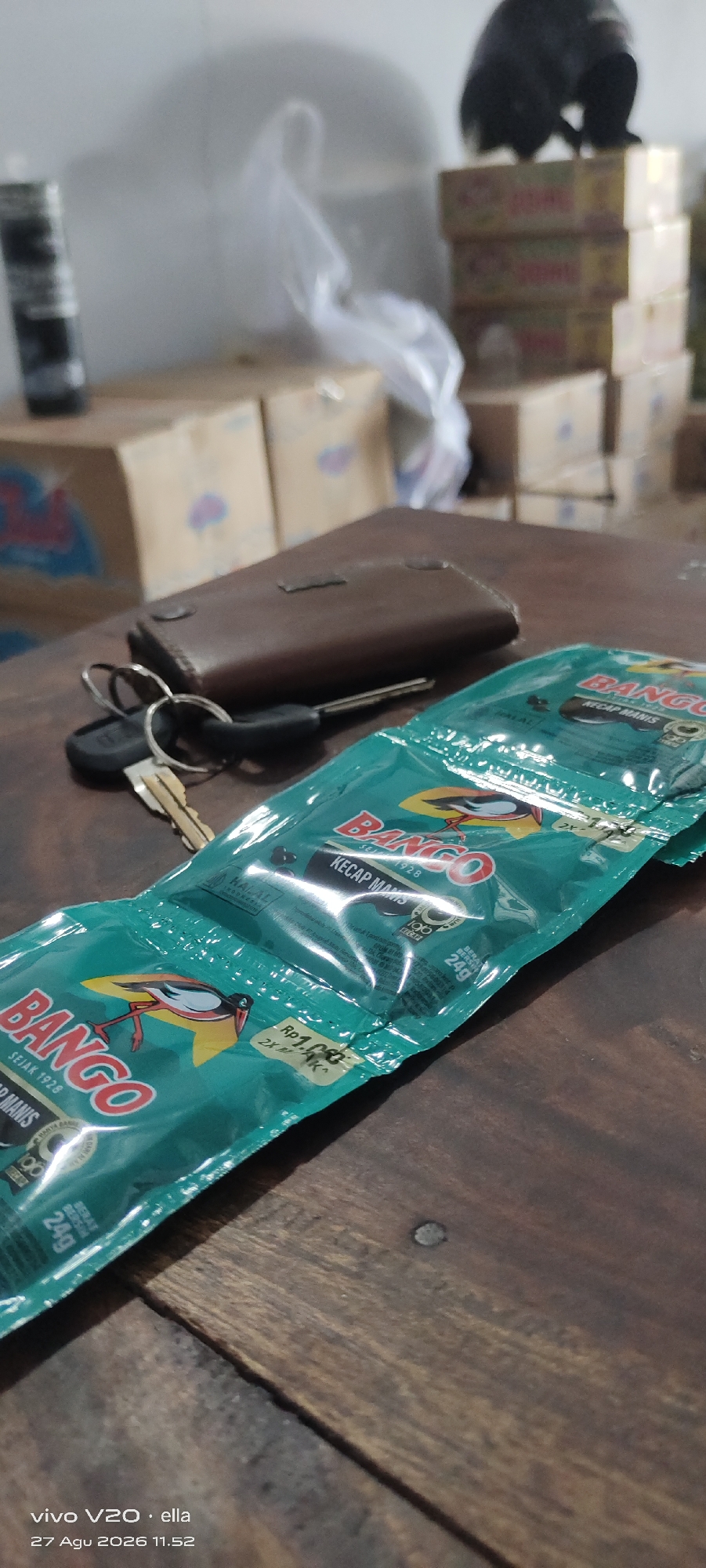 In the city of Simo, Indonesia, on a wooden table, two packets of Bango Kecap Manis are prominently displayed. To the left of the packets, a brown leather wallet and a set of keys are placed. In the background, stacks of cardboard boxes are visible, along with a blurry, dark object on top of one of the stacks. The lighting suggests an indoor environment, possibly a shop or storage area. The overall scene is a still life composition with everyday objects.