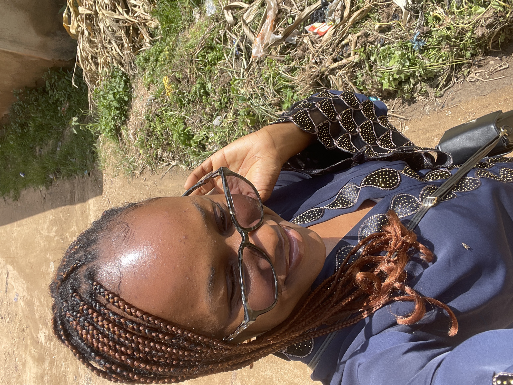 A young Black woman with braided brown hair and glasses poses for a selfie in what appears to be an outdoor, possibly rural or suburban, environment. She is wearing a dark blue garment with gold and black embellishments around the neckline and sleeves. A black purse is slung across her shoulder. Her right hand is raised to adjust her glasses, and she is smiling slightly. The background shows a dirt path to the left and overgrown vegetation, dried stalks, and some litter to the right. The lighting suggests it is daytime and sunny. The specific location in Rafin Par, Nigeria, is not discernible from the visual information in the image.