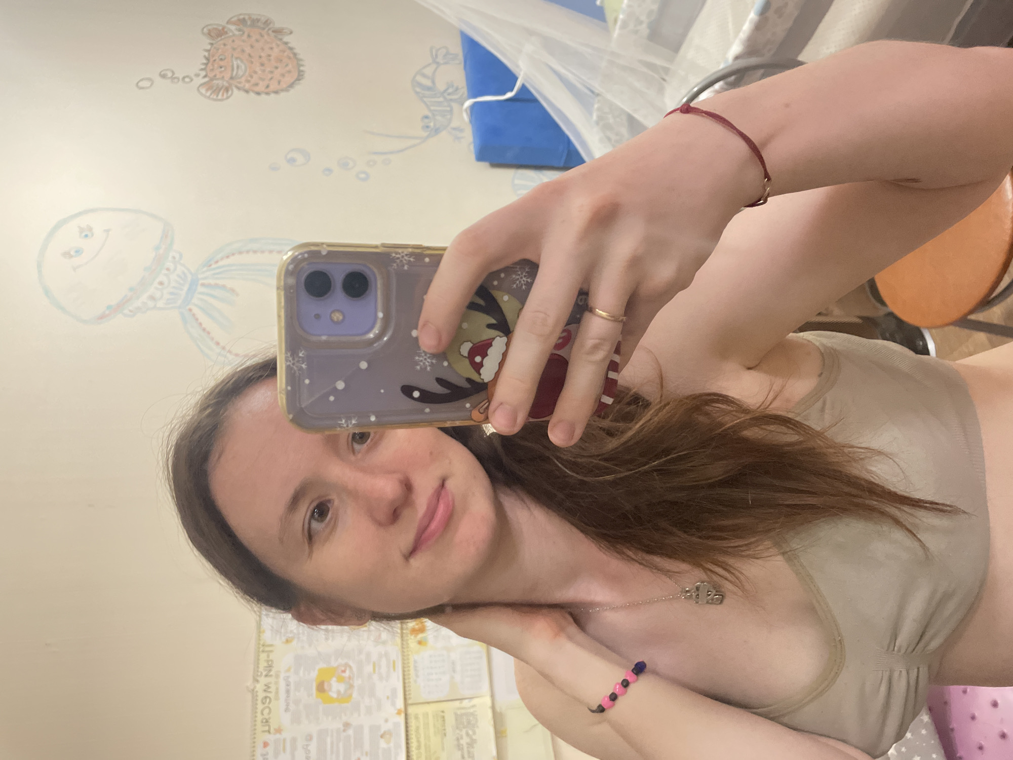 A woman is taking a selfie indoors. She is holding a smartphone with a decorated case. She is wearing a bra and a necklace. There is a red string bracelet on one wrist and a beaded bracelet on the other. The background shows a wall with painted sea creatures and a calendar or document. The location is Подстёпки, Russia.