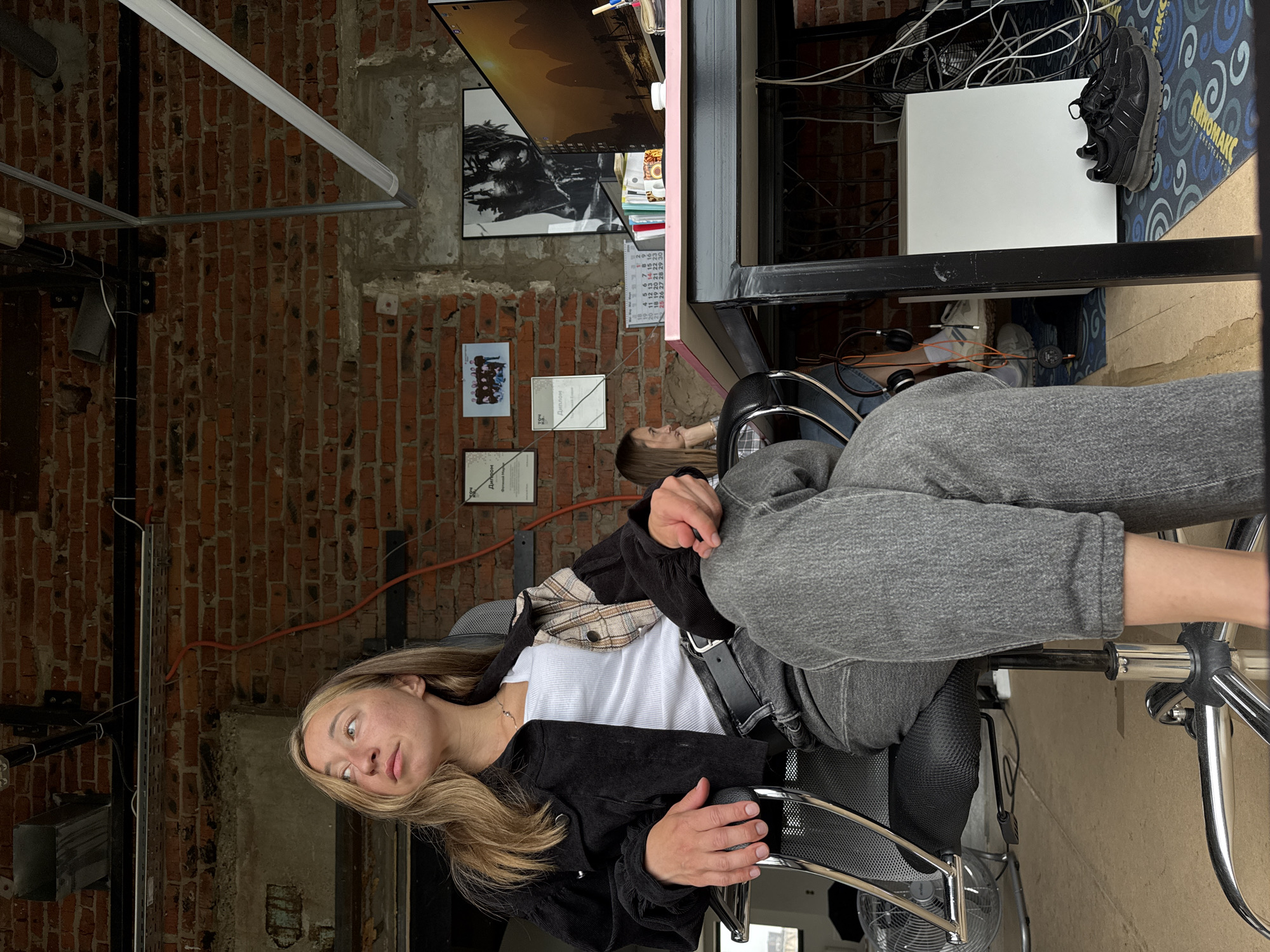 The image depicts an indoor scene, possibly an office or studio, with exposed brick walls and industrial-style lighting. In the foreground, a young woman with blonde hair is seated in an office chair, looking upwards and to her left. She is wearing a white tank top, a black corduroy jacket with plaid accents on one sleeve, and grey jeans. Her hands are resting on her lap.
In the mid-ground, another woman with blonde hair is visible sitting at a desk, looking towards the computer screen. The desk is pink and black, with various items on it, including books, a calendar, and a computer monitor displaying a landscape image. Behind the desk, artwork is displayed on the brick wall, including framed photographs and a large black and white poster.
The environment appears somewhat cluttered, with electrical cords and a fan visible in the background. The floor is covered with a patterned rug. The overall impression is a casual, creative workspace. There is no specific indicator within the image to suggest a location in Chaikovsky, Russia.