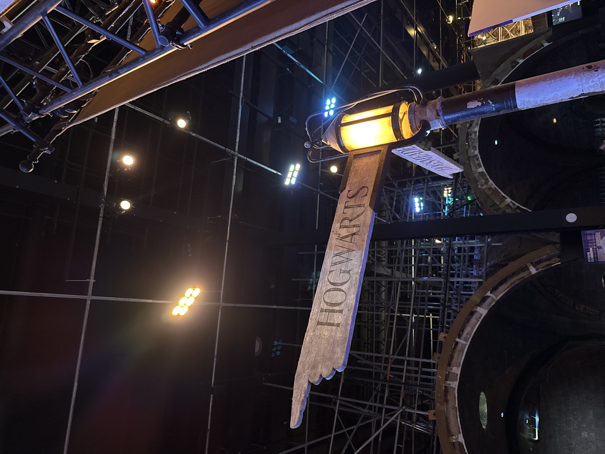 The image shows a wooden signpost with two directional signs, one pointing left and reading "HOGWARTS" and another pointing right, partially obscured, that reads "HOGSMEADE". The signpost is illuminated by an old-fashioned street lamp. In the background, there is scaffolding, large arched structures, and bright stage lighting. The setting appears to be an indoor event or exhibition. No specific location in the city of Three Rivers, United Kingdom can be determined from the image. There are no people clearly visible in the image.