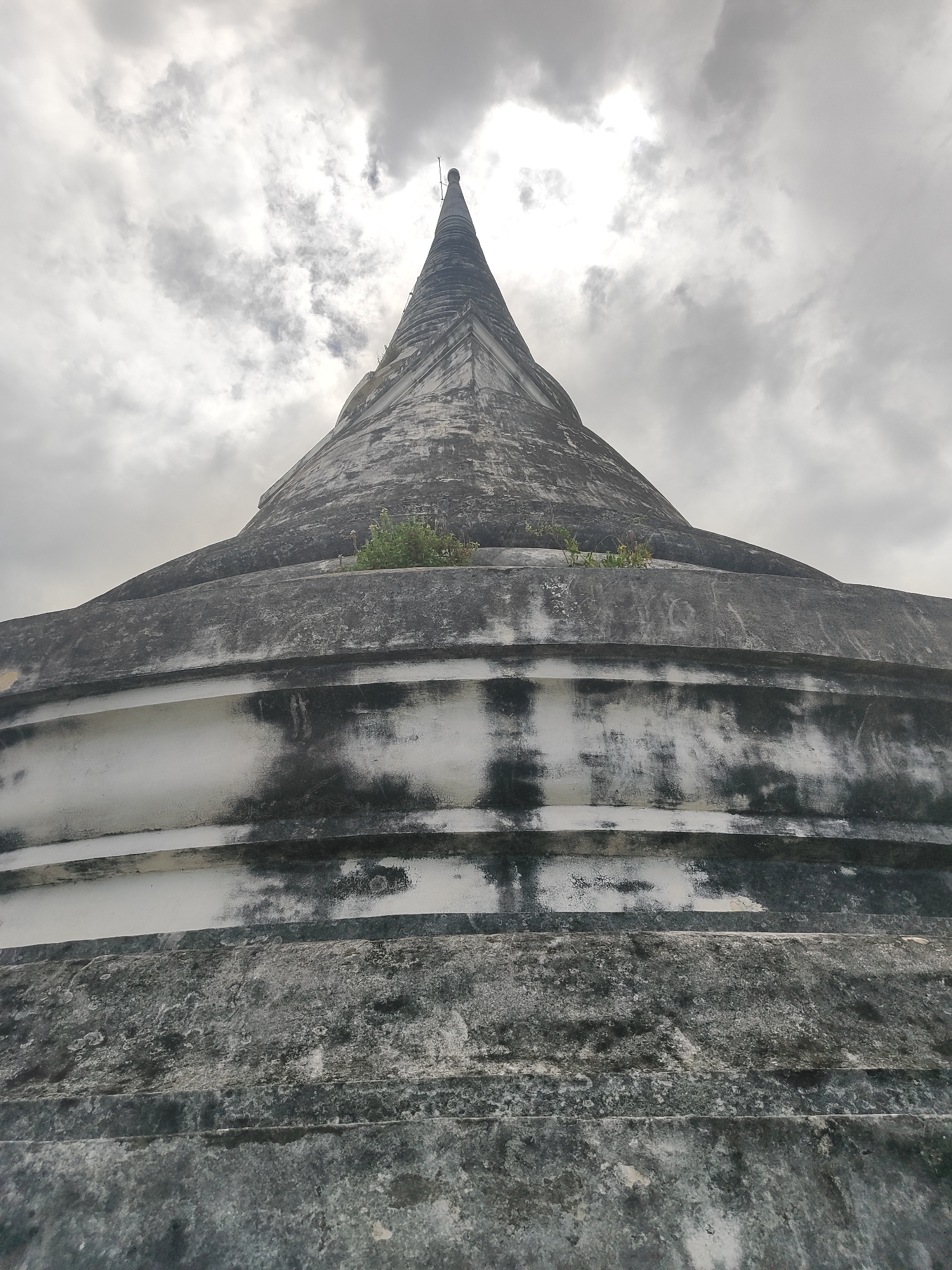 A low-angle, upward shot captures the upper portion of an ancient, weathered, conical stupa against a dramatic, overcast sky. The stupa, primarily rendered in shades of gray and white, exhibits significant signs of age, with patches of dark grime and what appears to be moss or lichen growing on its surfaces. The base of the stupa is broad and tiered, with distinct horizontal bands that decrease in width as they ascend. These bands are heavily textured and stained, suggesting long exposure to the elements. A sparse patch of green vegetation is visible growing from a crevice on one of the upper tiers. The apex of the stupa tapers to a sharp point, topped by a small finial. The sky is filled with heavy, gray clouds, with a brighter area of diffused light suggesting the sun is behind them, creating a somber and atmospheric backdrop. The image appears to be taken at Wat Mahathat Worawihan in Phetchaburi, Thailand, an important historical temple known for its ancient architecture. There are no people visible in the image.
