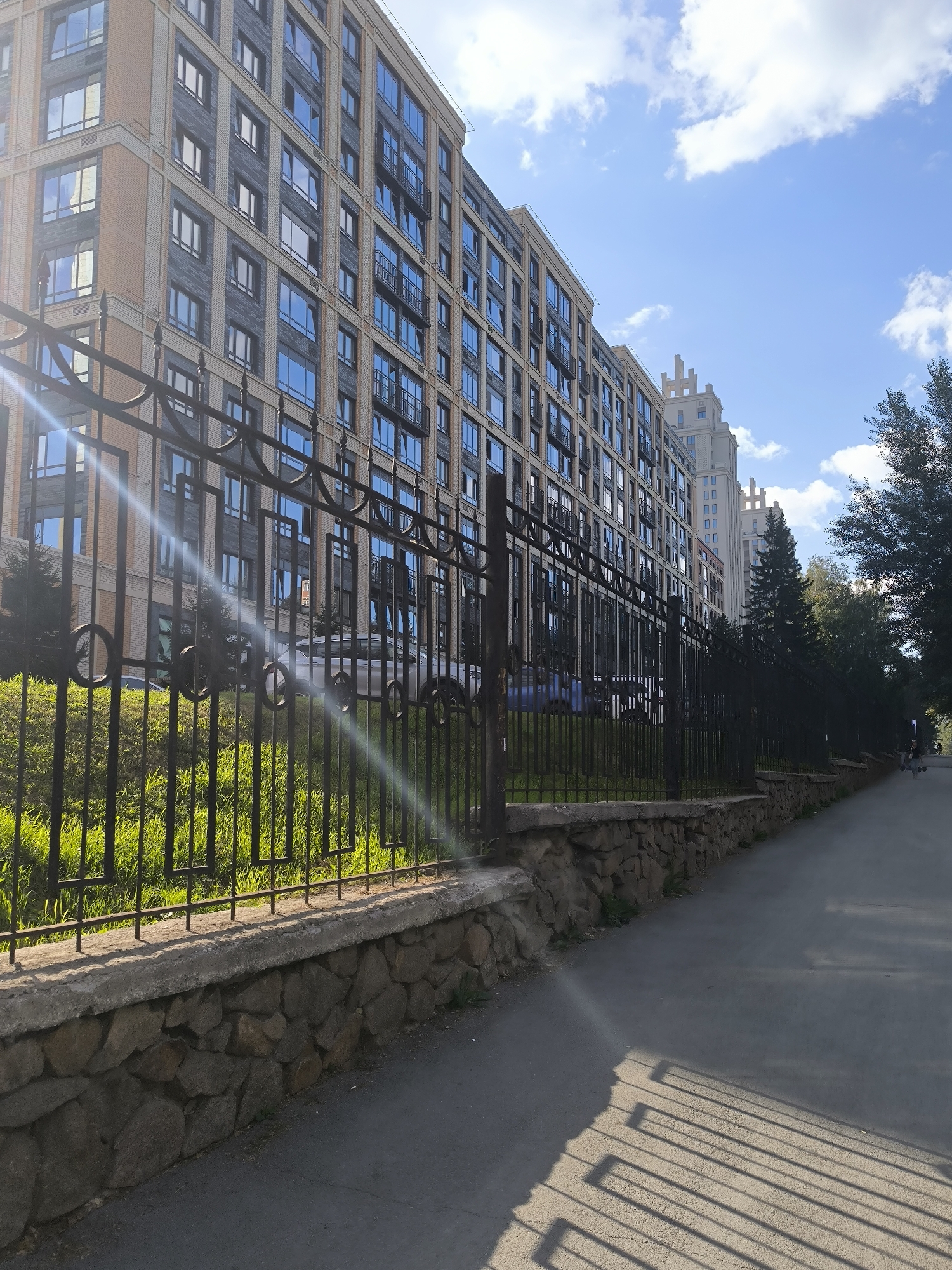 A sun-drenched street in Novosibirsk, Russia, runs alongside a modern, multi-story building with beige brick and dark gray stone accents, featuring numerous blue-tinted windows and balconies. A black wrought-iron fence, topped with decorative elements, separates the building's green lawn from the paved sidewalk. The fence is supported by a stone wall. Further down the street, other similar buildings are visible, along with trees and a few individuals walking. Sunlight casts long, distinct shadows of the fence across the sidewalk, creating a geometric pattern. The sky above is blue with scattered white clouds.