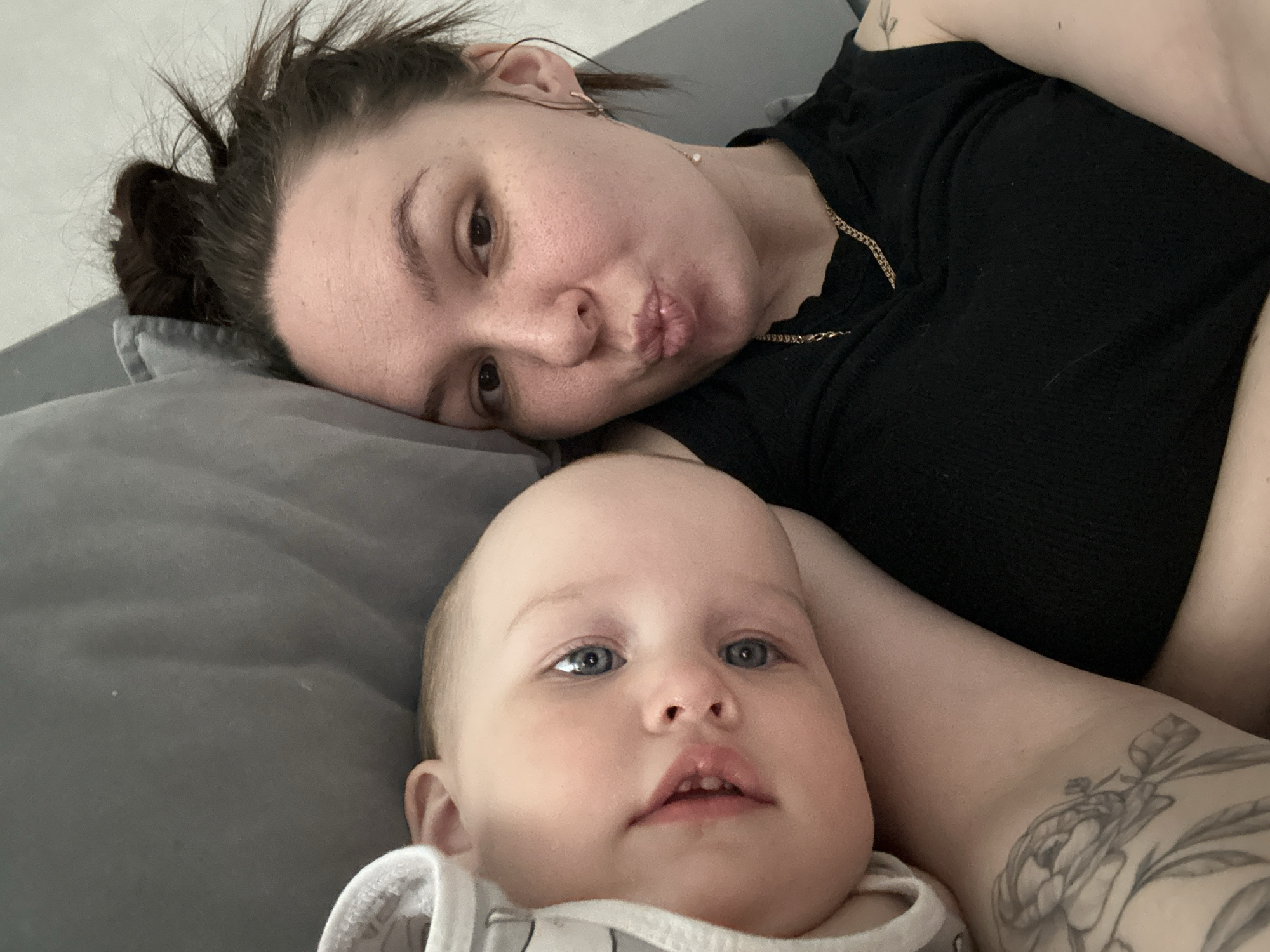 An intimate, close-up shot features a woman and a baby lying on a grey surface, likely a bed or sofa. The woman, positioned in the upper part of the frame, has dark hair styled in a messy bun. She looks directly at the camera with a playful expression, her lips puckered. She wears a black ribbed top and a gold chain necklace. Her right arm, with a prominent floral tattoo, is visible in the foreground, supporting her or the baby.
The baby, positioned below the woman, has light hair and striking blue eyes, also looking towards the camera. Its mouth is slightly open, revealing two small bottom teeth. The baby is wearing a white garment with subtle patterns or stripes. The overall scene conveys a tender and relaxed moment between mother and child, possibly a playful selfie or a moment of rest. No specific time of day or weather is discernible from the indoor setting. There are no visible texts in the image. The provided location context is Chaikovsky, Russia.