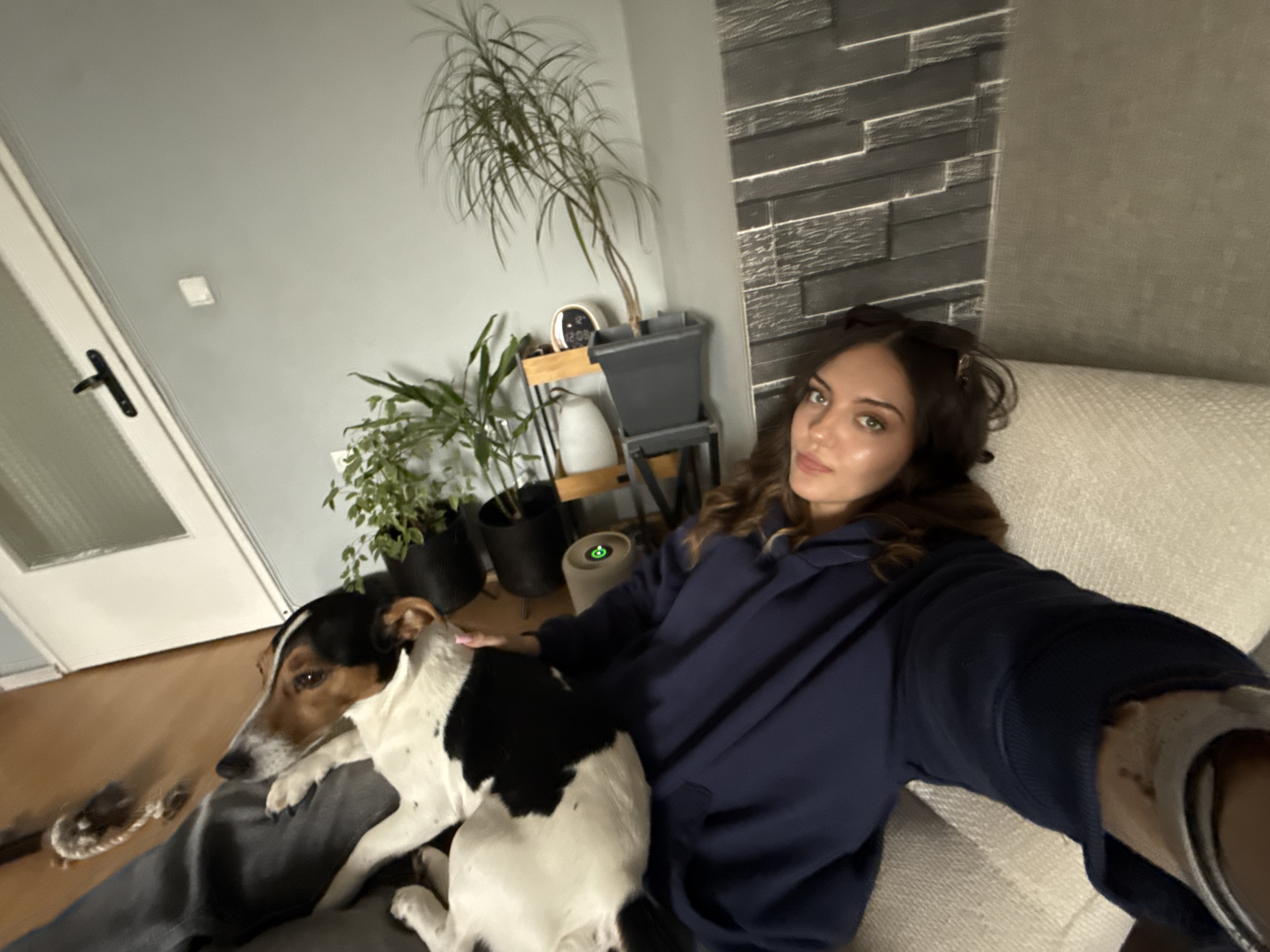 A woman is sitting on a couch, holding a dog. The dog is lying on the woman’s lap. There are several plants in the background. The interior suggests a home in Sofia, Bulgaria.
