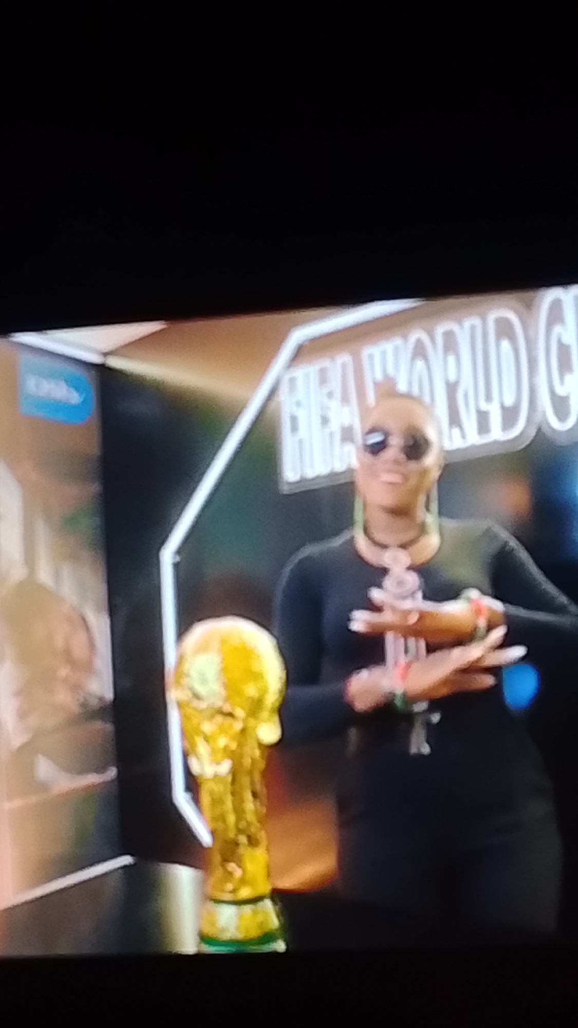 The image shows a woman wearing sunglasses, a black top, and dark pants, standing next to the FIFA World Cup trophy. She is wearing a beaded necklace and bracelets on her wrists. Her hands are clasped in front of her. The trophy is golden and appears to be on a stand. Behind them, a large sign reads "FIFA WORLD CUP". The scene is likely a promotional event. The city of Lokoja, Nigeria is not mentioned in the image.