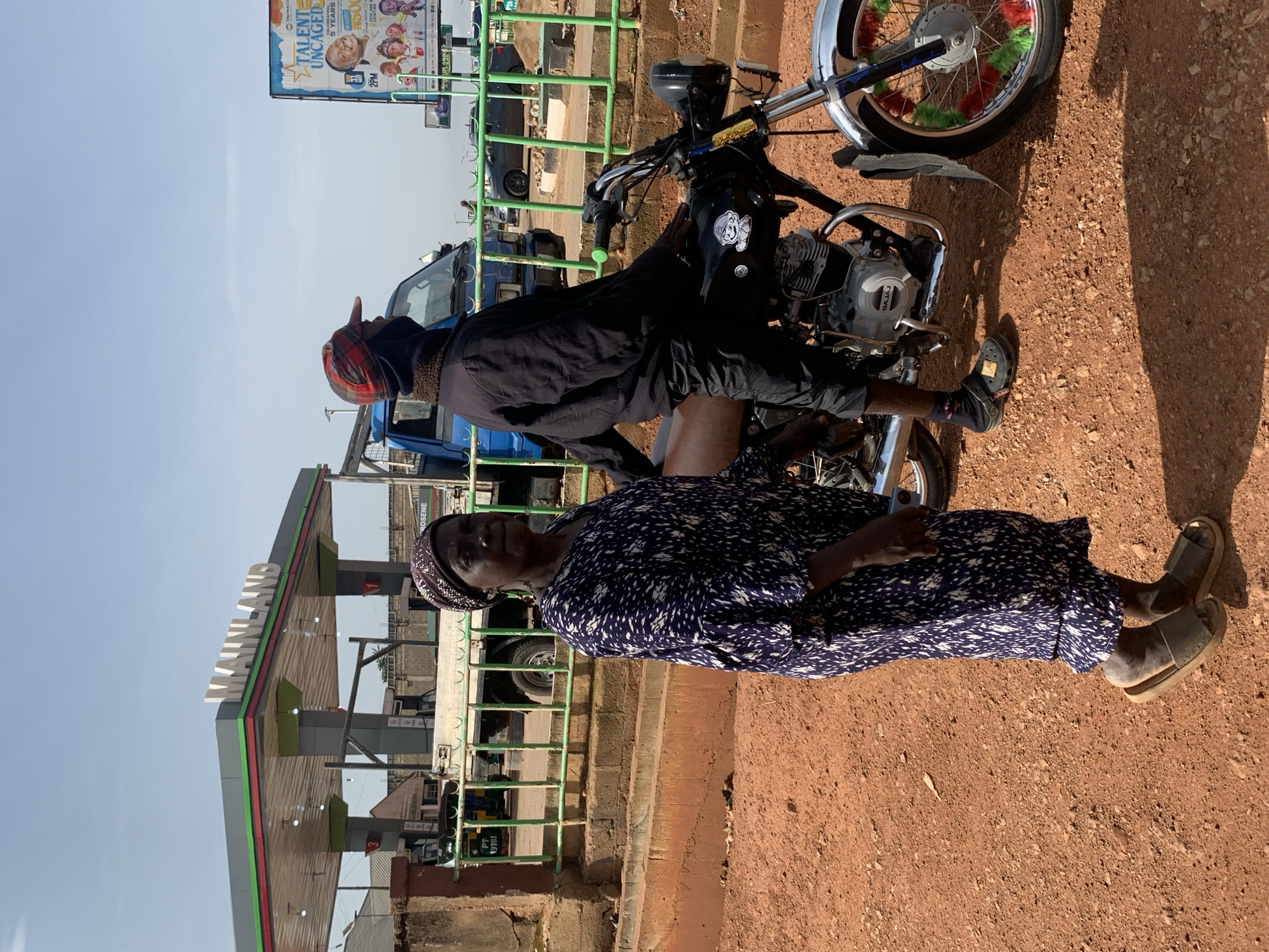 In Gura Top, Nigeria, a woman in a blue and white patterned dress stands in front of a motorcycle. She is wearing a headscarf and sandals. A man wearing a red and black checkered cap and a black jacket is sitting on the motorcycle. In the background, there is a gas station with the name "MAINLAND" visible. A blue truck is parked behind a green metal fence. A billboard with the words "TALENT UNCAGED" is also visible. The ground is covered in reddish-brown dirt.