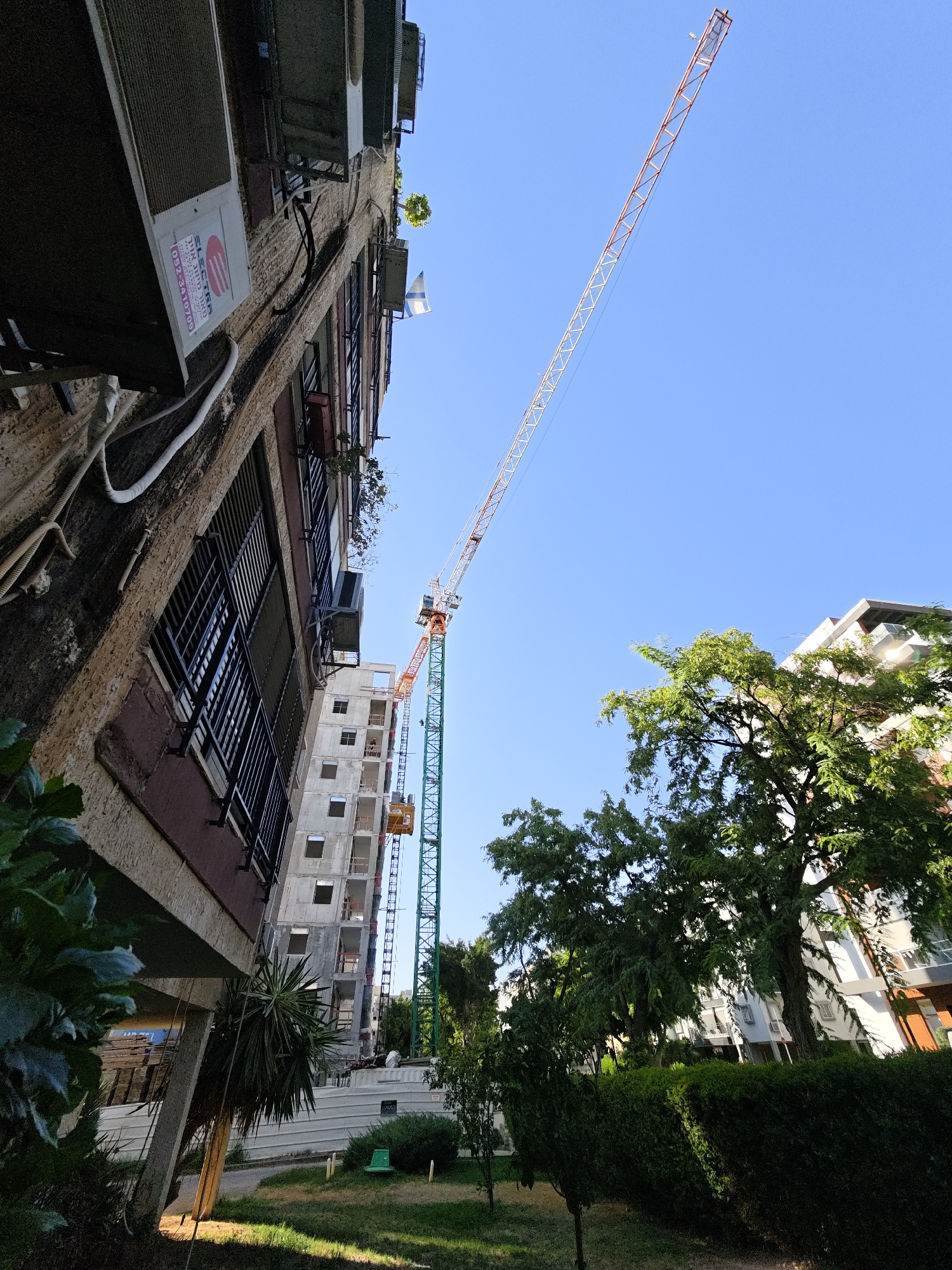 In Petah Tikva, Israel, a construction site is visible, featuring a tall crane with an orange and white boom extending across a clear blue sky. Adjacent to the crane is a multi-story building under construction, with visible scaffolding and concrete structures. In the foreground, a building with balconies and air conditioning units is seen from a low angle. The scene includes lush green trees and manicured bushes, suggesting an urban outdoor environment. The image appears to be taken during daylight hours, with shadows cast by the sun.