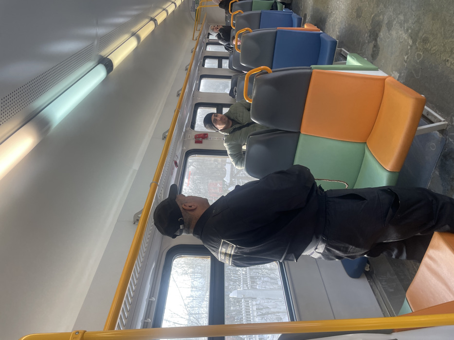 The image displays the interior of a commuter train car, specifically located in Fryazevo, Russia, during daylight hours. A man in a black uniform, including a black baseball cap and a jacket featuring a light-colored rectangular patch with Cyrillic text on the upper back, stands with his back partially to the viewer, facing another man. A string of reddish-brown beads hangs from the uniformed man's left side. The second man wears a dark beanie and an olive-green jacket, and is positioned further down the aisle, appearing to lean against a seat while looking towards the uniformed individual.
The train car features a white ceiling equipped with multiple fluorescent tube lights, some emitting a cool white light and others a warmer yellow light. Yellow grab rails are mounted above the windows, through which a snow-covered exterior landscape with trees and a white fence is visible. The seating consists of rows of multi-colored upholstered seats, featuring segments of green, orange, blue, and dark gray. A red emergency call button is mounted on the wall between two windows. Several other passengers are partially discernible further along the aisle in the background. The floor appears to be a dark, textured surface.