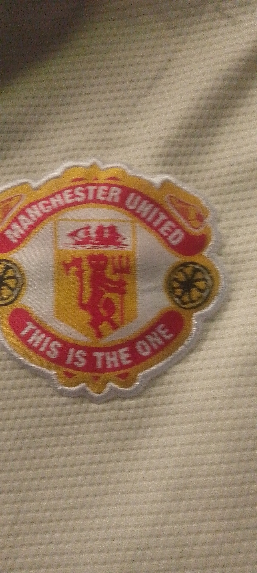 The image shows a close-up of a Manchester United football club crest, embroidered on a textured fabric, likely a piece of clothing or merchandise. The crest features a red devil in the center, a ship above it, and a red banner with the words "MANCHESTER UNITED" arching over the top. Below the central shield, another red banner displays the text "THIS IS THE ONE" in white lettering. The crest is predominantly red, yellow, and white, with black accents on some of the details. The fabric it is on appears to be a light beige or cream color with a visible woven pattern. The lighting is somewhat dim, casting a slight shadow. There are no people, specific landmarks, or environmental details visible to determine a location in Jalingo, Nigeria.