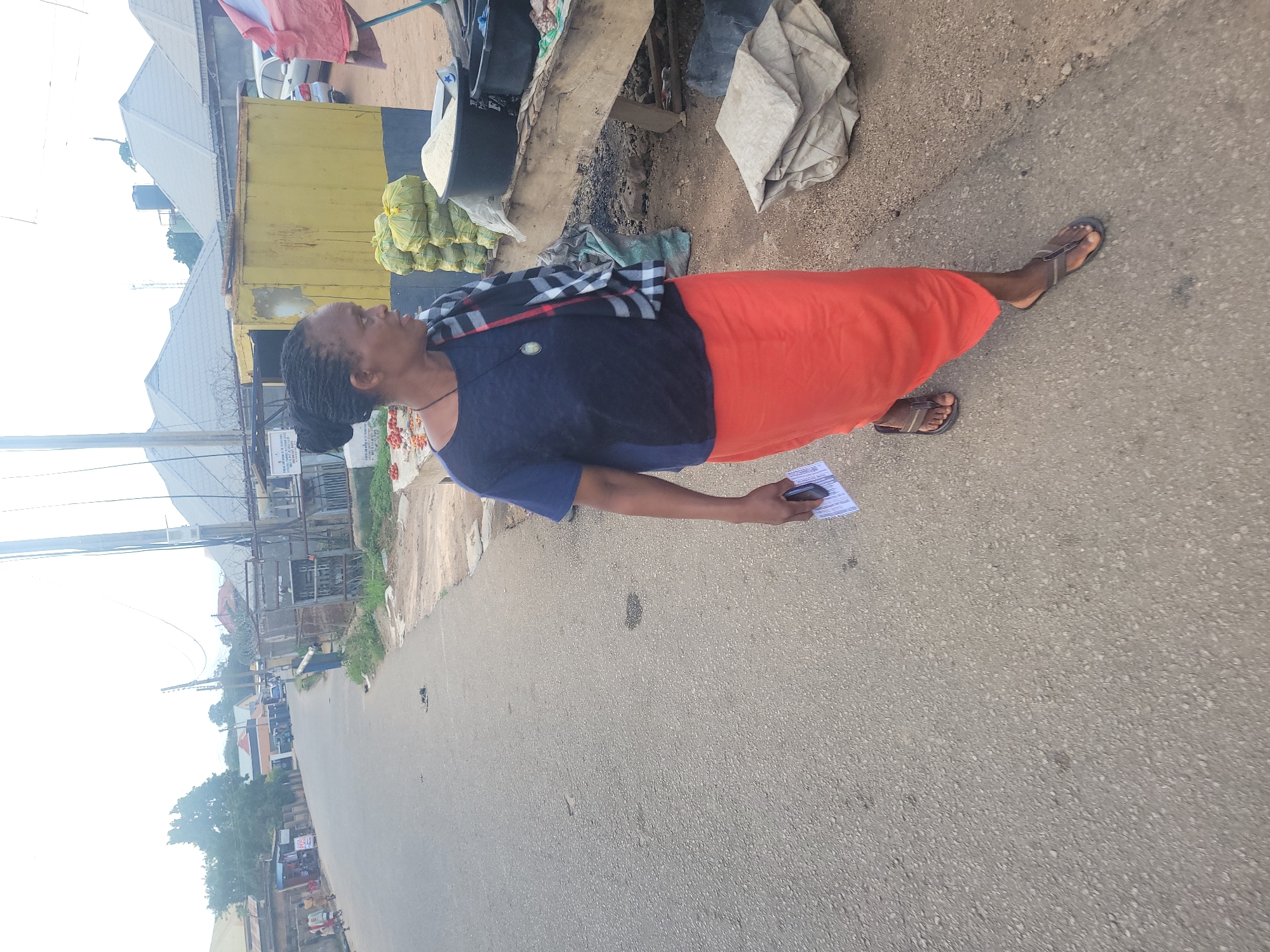 A woman walks on an asphalt road in Jos, Nigeria. She has dark skin, braided hair pulled back, and wears a dark blue t-shirt and an orange skirt. Around her neck, she wears a black and white checkered scarf with a pendant. She is holding a white card in her right hand. To her right, a yellow kiosk with a faded blue stripe is visible. Stacks of green-bagged produce and a large black container filled with white grain are on a wooden table in front of the kiosk. In the background, other buildings, power lines, and a street with a few vehicles and pedestrians can be seen. The scene appears to be a marketplace.