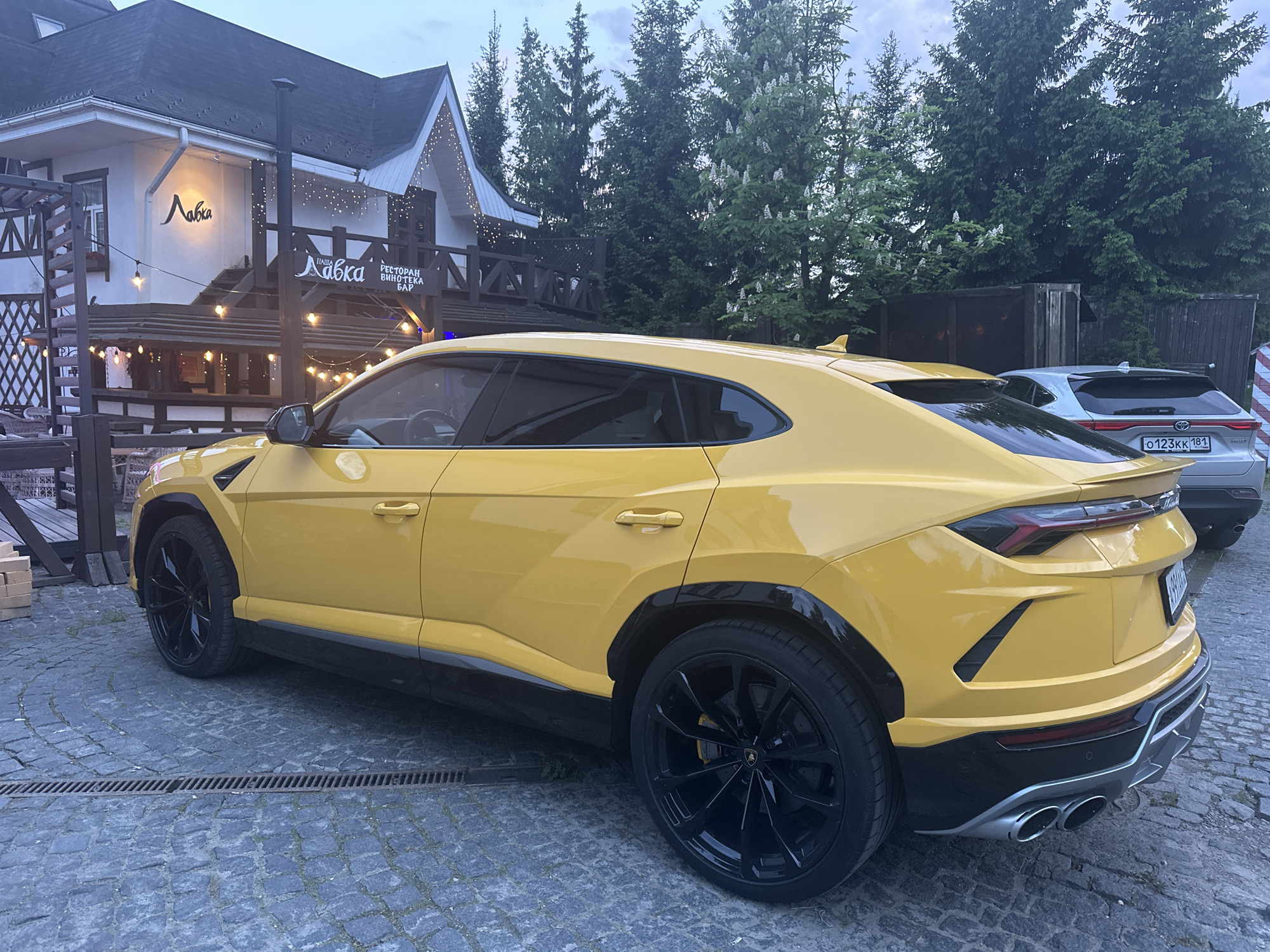 A yellow Lamborghini Urus is parked on a cobblestone road in front of a building with a sign reading "Лавка" and "РЕСТОРАН ВИНОТЕКА БАР" (Restaurant, Winery, Bar). Another car is partially visible behind the Lamborghini, displaying the license plate "O123KK 181". Tall evergreen trees are in the background, and the scene appears to be located in Новоглаголево, Russia.