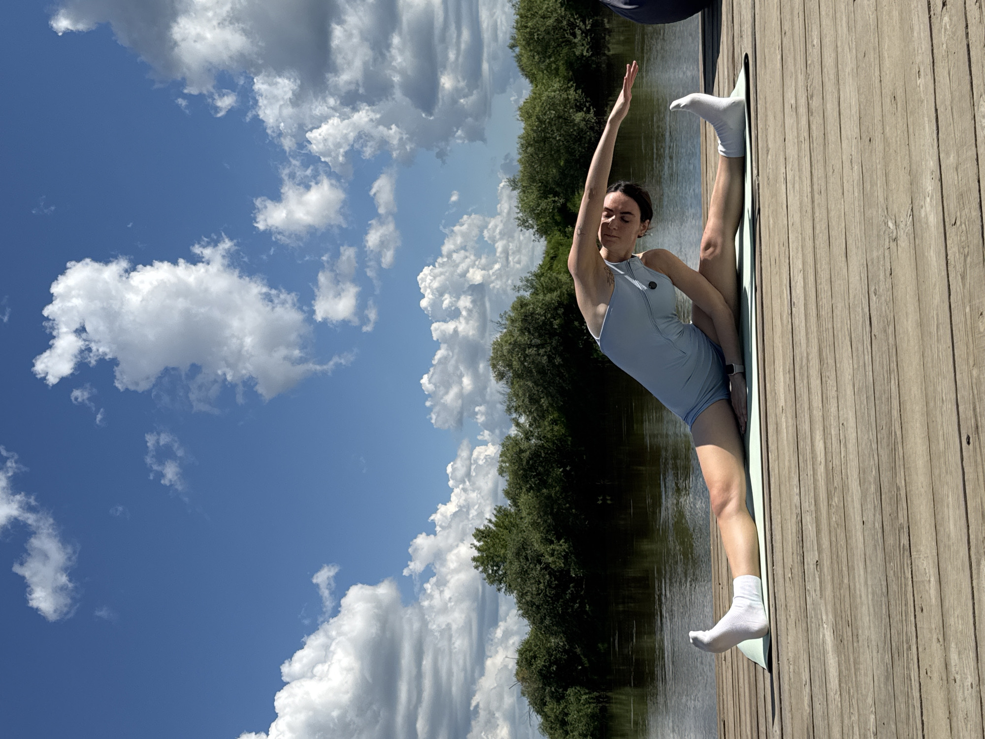 A woman is performing a yoga pose on a wooden pier next to a body of water. She is wearing a light blue athletic outfit and white socks. Her left leg is extended forward, and her right leg is bent with her foot near her inner thigh. She is leaning to her right, with her right arm extended overhead. The pier is made of weathered wooden planks. In the background, there is a calm body of water, with lush green trees and bushes lining the opposite bank. The sky above is a vibrant blue with scattered white cumulus clouds. The scene appears to be set in the city of Онохино, Russia, given the provided context.