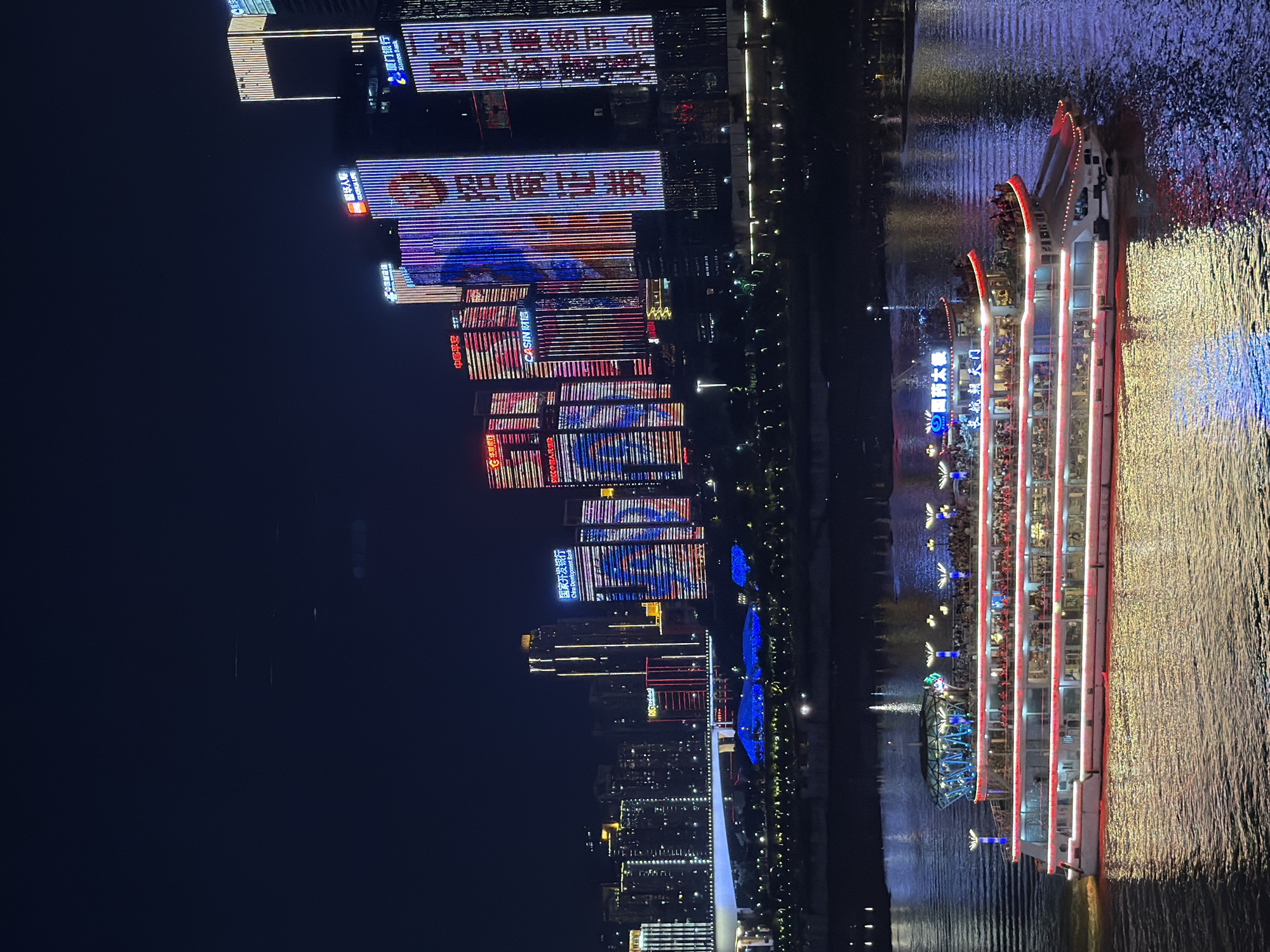 This is a nighttime, low-angle shot of a city skyline and a river. The city is illuminated with bright lights from buildings, some featuring colorful LED displays. Visible text on the buildings includes "国家开发银行" (China Development Bank), "中国平安" (Ping An Insurance), "CASIN 财信" (Caisin), "国药太极" (Guoyao Taiji), "长航朝天门" (Changhang Chaotianmen), "国华人寿" (Guhuaren Life), "中国邮" (China Post), "招商证券" (China Merchants Securities), and "厦门银行" (Xiamen Bank). The river in the foreground reflects the lights of the city and is traversed by a multi-tiered illuminated boat filled with people. The boat has red and white lights outlining its decks, and the water is rippled, creating a shimmering effect. The overall scene suggests a vibrant urban environment at night, likely a tourist area or waterfront promenade. The time of day is clearly night, and the weather appears clear, allowing for the vibrant light displays.