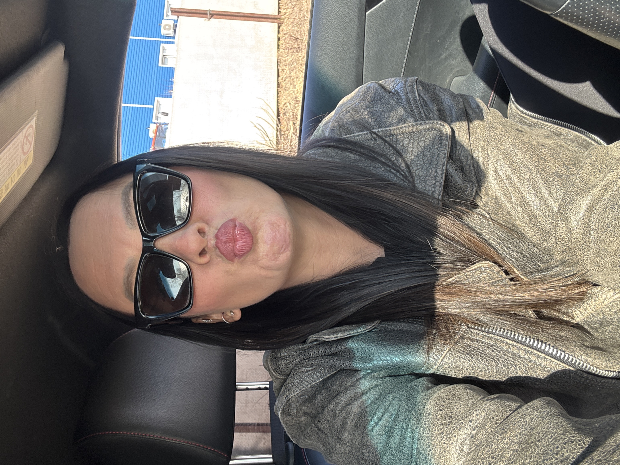 A woman is in a car wearing sunglasses, a grey leather jacket, with black hair. In the background through the window is a building. Given the prompt, the location is likely Chaikovsky, Russia.