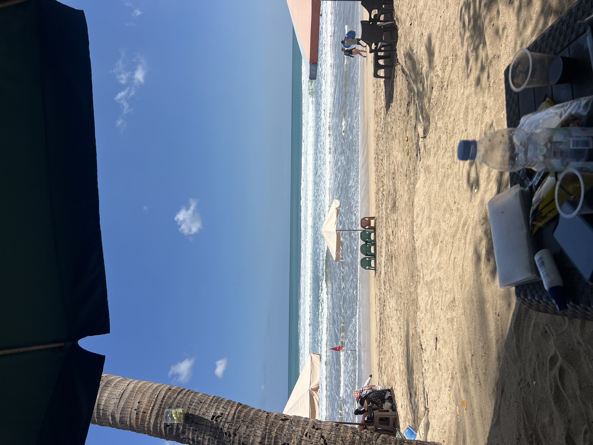 A sunny day at Kuta Beach, Indonesia. The image captures a view of the ocean with moderate waves crashing on the sandy shore. In the foreground, a table with a water bottle, plastic cups, and a phone is visible, suggesting a relaxed setting for beachgoers. Several beach umbrellas and chairs are scattered on the sand, some occupied by individuals. Two people are seen walking on the beach carrying surfboards, indicating the presence of water sports enthusiasts. The scene is framed by the leaves of a palm tree on the left and a large beach umbrella on the right, contributing to the tropical ambiance of the location. The sky is clear blue with a few scattered clouds.