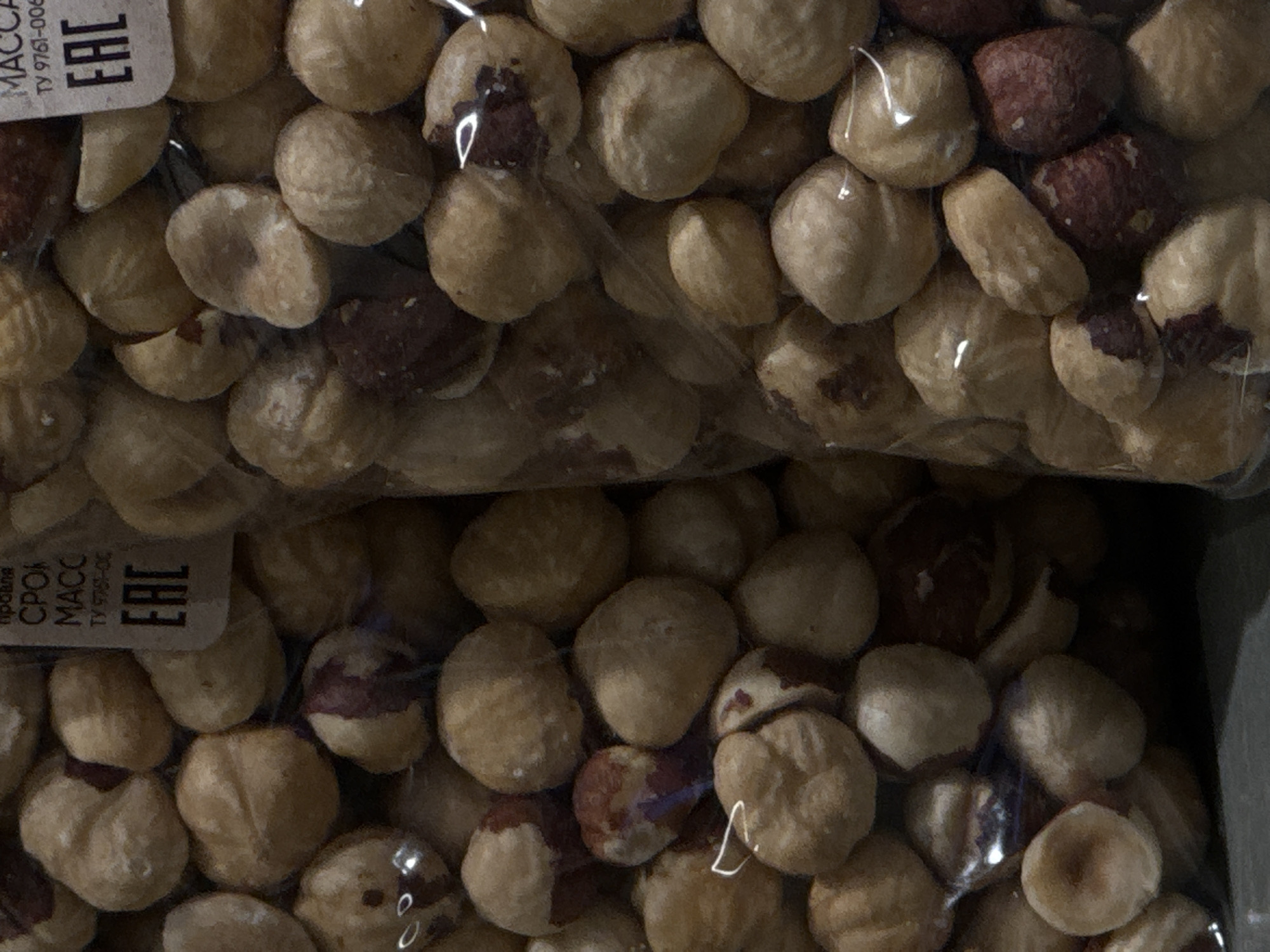 The image is a close-up, vertical shot of a package of hazelnuts. The nuts are light brown and some have reddish-brown skins. They are densely packed within clear plastic packaging. Text is visible on a label on the right side of the packaging, including the letters "EAC" in a black rectangle. The label also appears to have Russian text, possibly indicating product information. The lighting suggests an indoor environment, and the focus is on the texture and detail of the hazelnuts. There are no people or discernible environmental cues that would allow for a specific location within Moscow to be identified.
