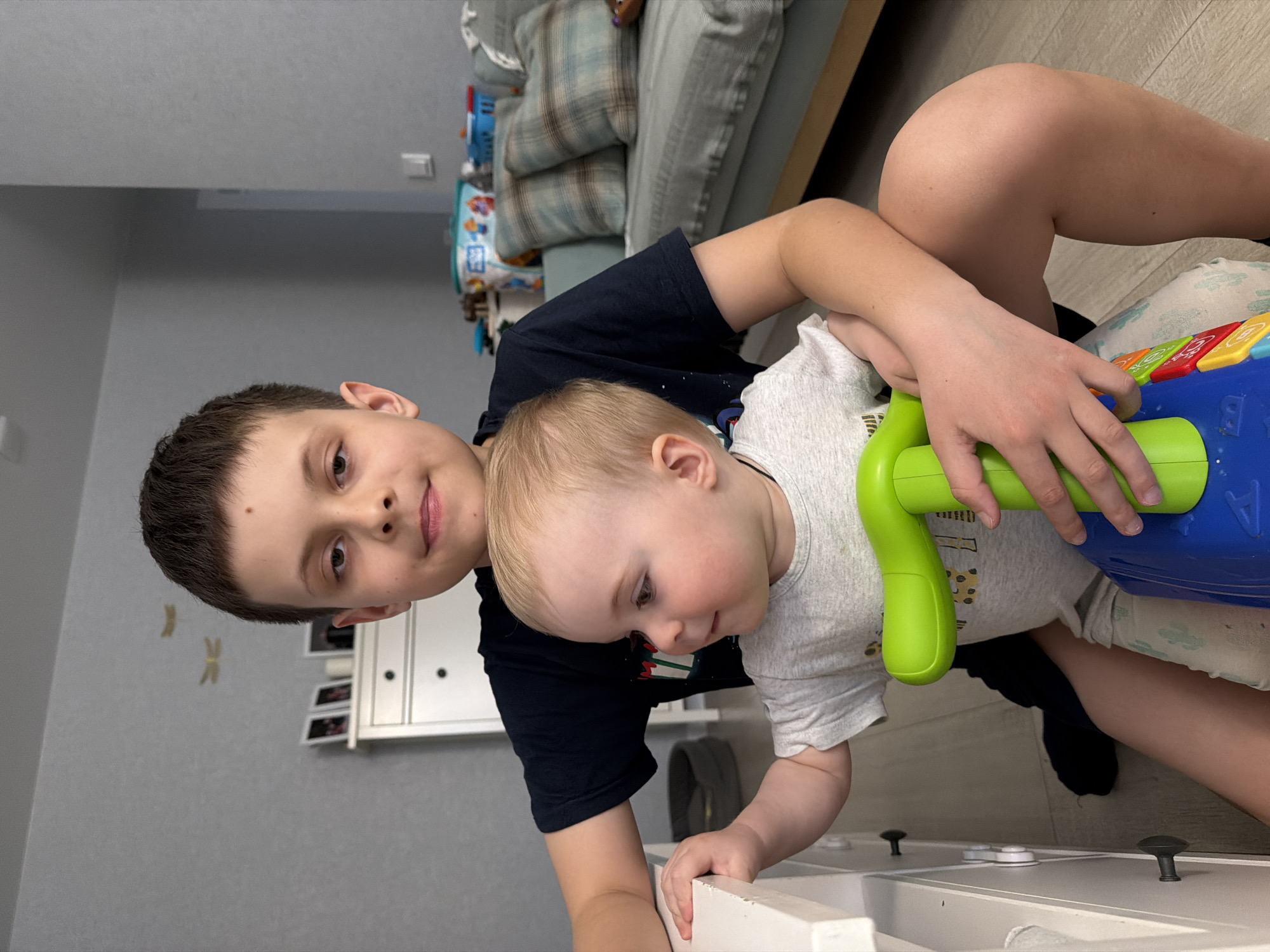 Two boys are playing indoors. The older boy is wearing a dark blue shirt. The younger boy has blond hair and is wearing a gray shirt. They are interacting with a blue and green toy while standing in front of a white dresser. The environment includes a gray wall, and a couch in the background. The location in the city of Votkinsk, Russia, cannot be confirmed.